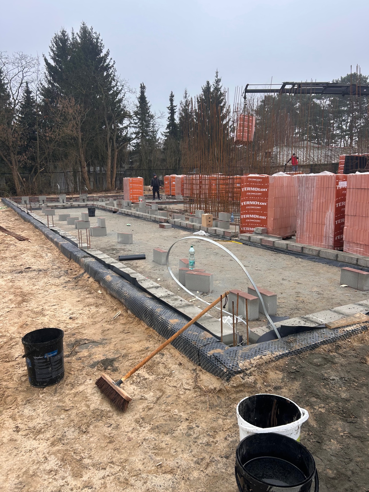 Fundamenty domu w trakcie budowy: widoczne bloczki fundamentowe, izolacja, zbrojenie i palety z cegłami. Na pierwszym planie wiadro i miotła.