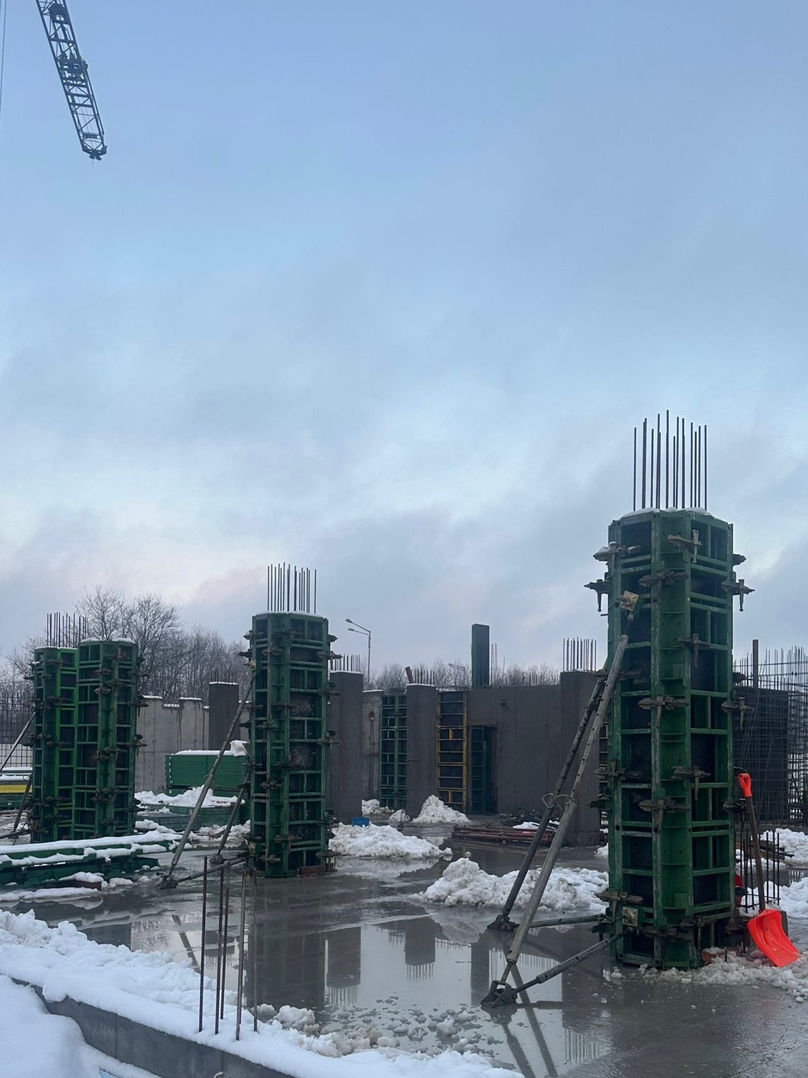 W zimowej scenerii, widok na plac budowy z zielonymi szalunkami kolumn i wystającymi prętami zbrojeniowymi, częściowo pokryty śniegiem i wodą. W tle widoczne elementy konstrukcji.