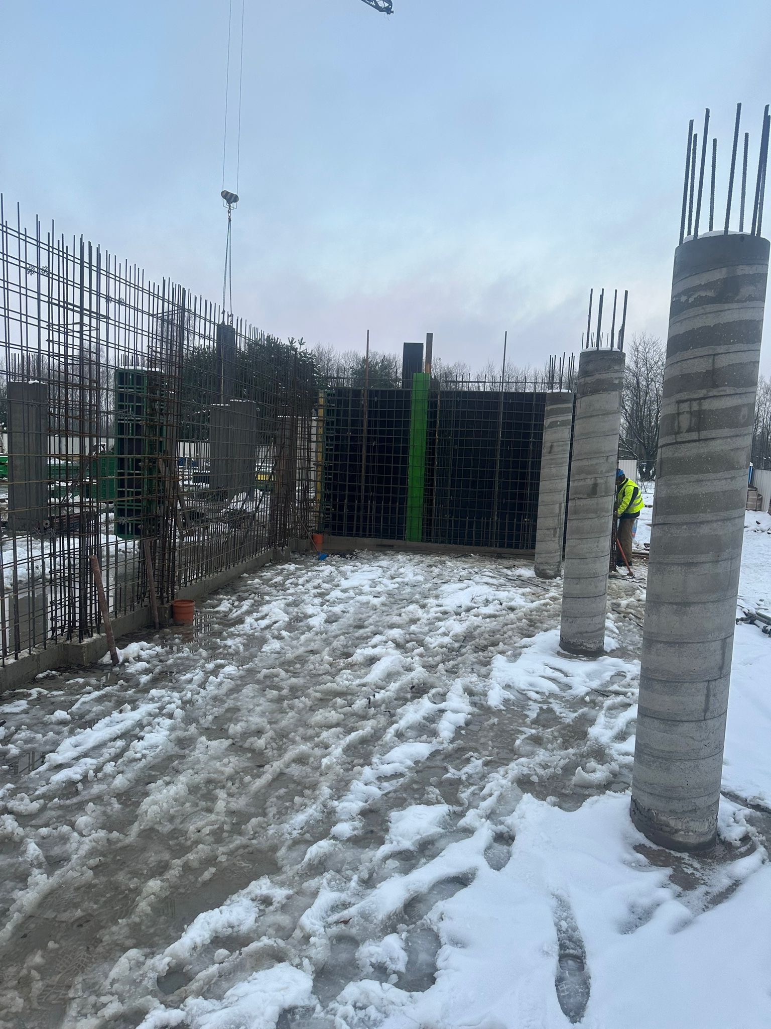 Zimowy plac budowy z widocznymi zbrojeniami ścian i kolumn. W tle zielone drzewa i pochmurne niebo. Pracownik w odblaskowej kamizelce przy kolumnie.