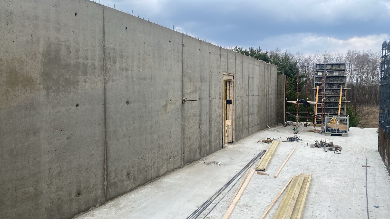 Surowa, betonowa ściana z otworem drzwiowym w trakcie budowy. Widoczne szalunki, zbrojenia i drewniane elementy. Szary beton kontrastuje z zielenią drzew w tle.