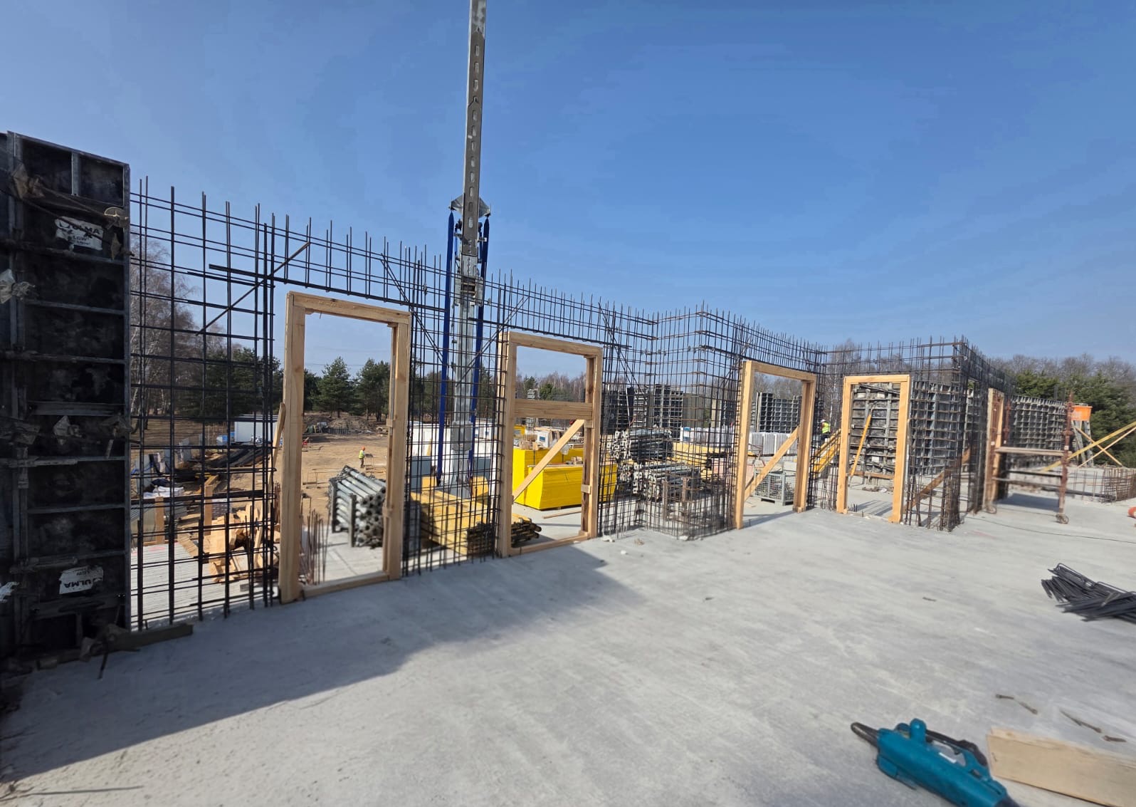 Zbrojenie fundamentów z otworami na drzwi, widok z góry na betonową płytę, w tle żuraw budowlany i drzewa. Słoneczny dzień, niebieskie niebo.