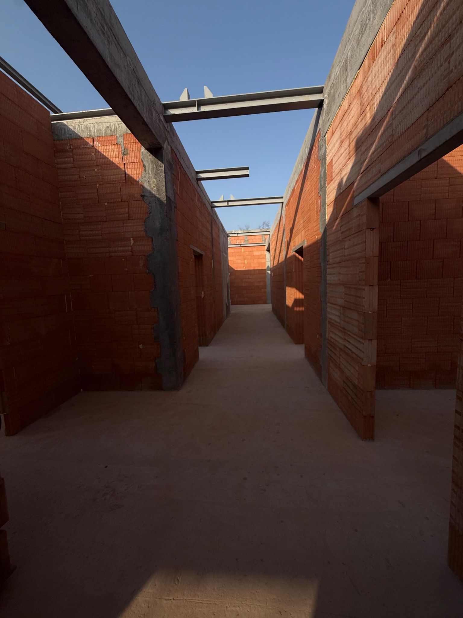 Wnętrze budynku w trakcie budowy. Widoczne ściany z czerwonej cegły, betonowe nadproża i stalowe belki stropowe. Surowy stan, przygotowanie do dalszych prac wykończeniowych.