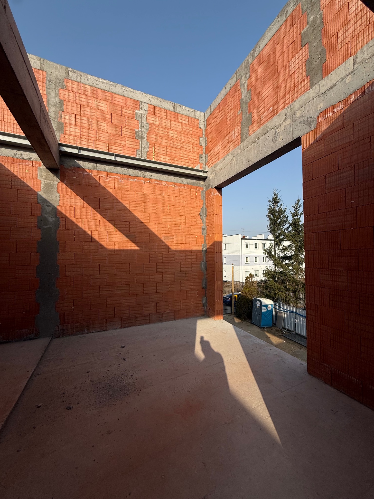 Wnętrze budynku w trakcie murowania ścian z czerwonej cegły. Widoczne betonowe nadproża i fragment nieba w tle. Na zewnątrz budynku widoczna toaleta przenośna.