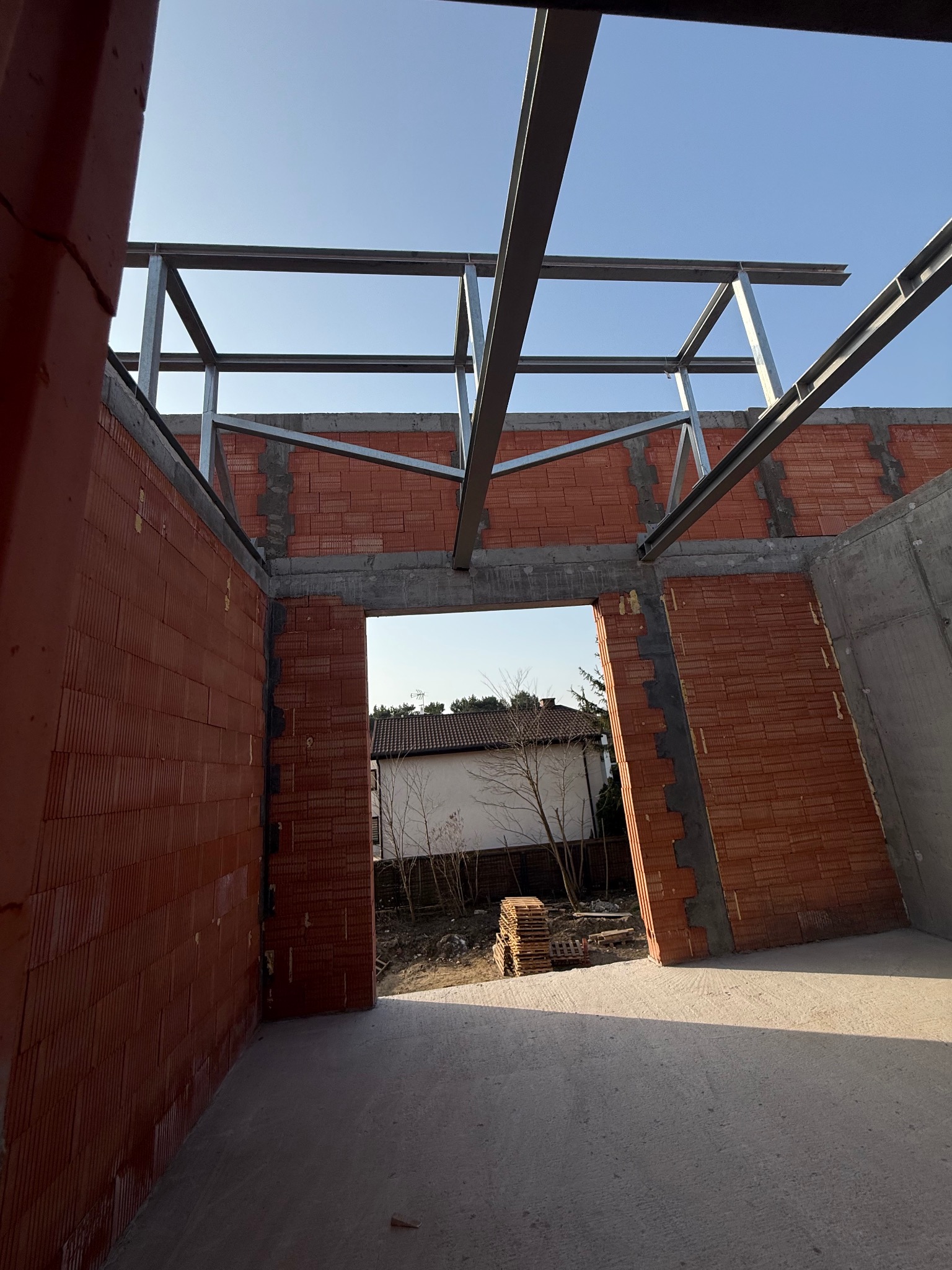 Wnętrze budynku w trakcie budowy: ściany z czerwonej cegły, betonowe elementy konstrukcyjne i stalowa rama pod dachem. Widok przez otwór drzwiowy na zewnątrz.