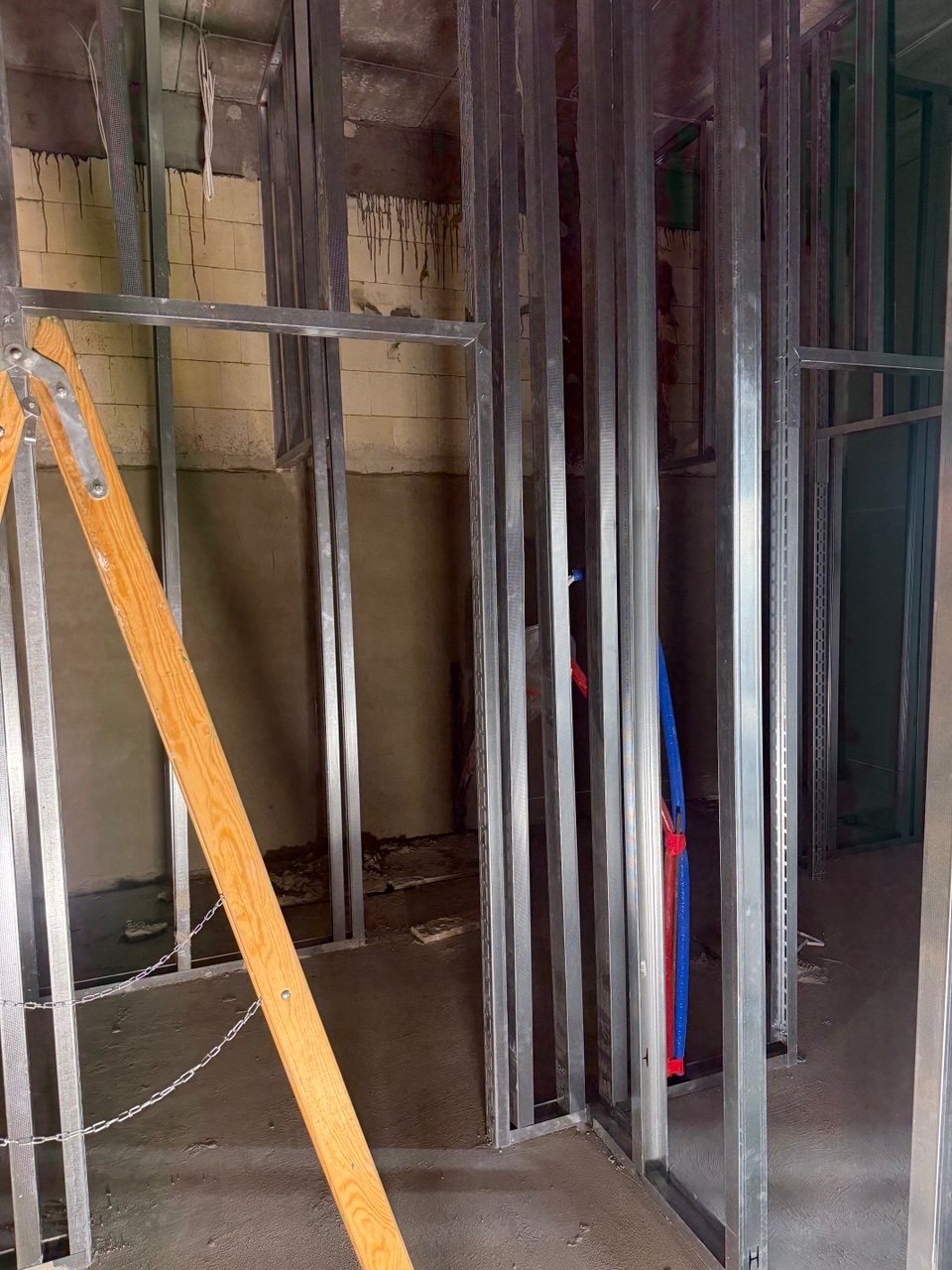 Wnętrze w trakcie budowy: metalowa konstrukcja szkieletowa ścian, widoczne instalacje (rury), fragment drabiny drewnianej. Surowy stan, przygotowanie do dalszych prac wykończeniowych.