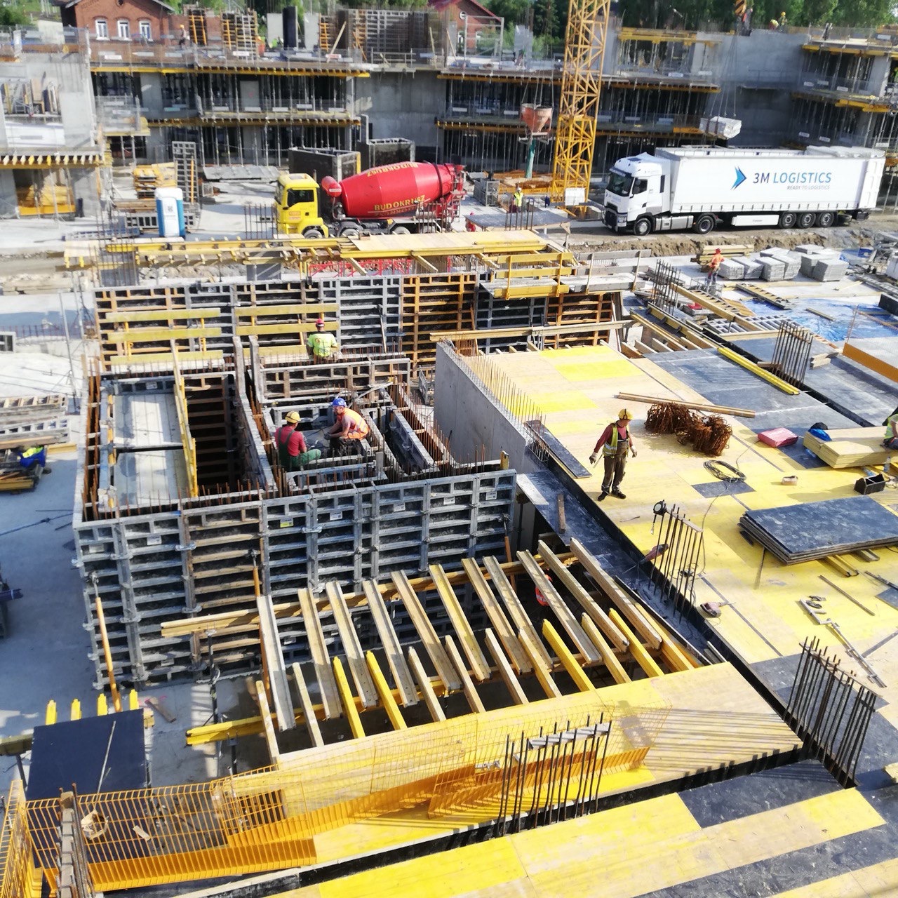 Wysoka perspektywa na plac budowy z pracownikami w szalunkach, betoniarką i ciężarówką logistyczną. Żółte elementy konstrukcyjne i zbrojenia w centrum kadru.