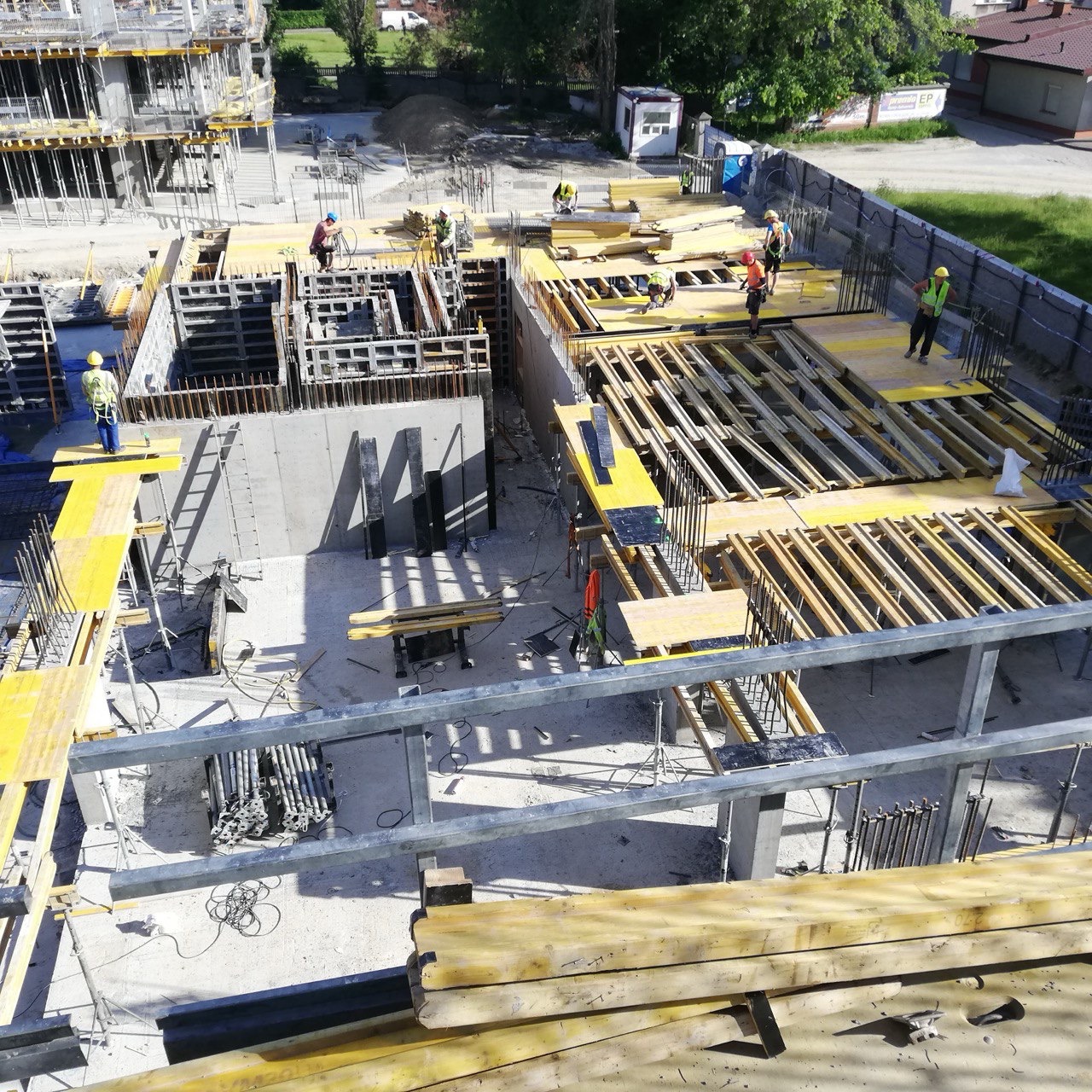 Wysoka perspektywa na plac budowy z pracownikami w kaskach, widoczne szalunki, zbrojenia i elementy konstrukcyjne. Jasne oświetlenie podkreśla detale budowy.