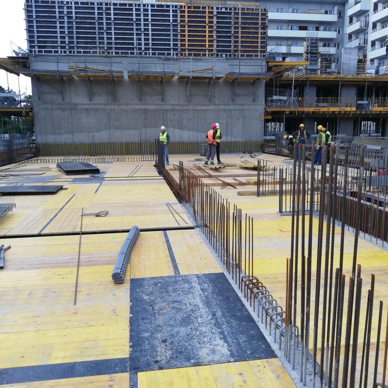 Wysoki kąt na plac budowy z widoczną konstrukcją żelbetową, deskowaniem i pracownikami w kaskach. Widoczne zbrojenia i elementy konstrukcyjne budynku w trakcie budowy.