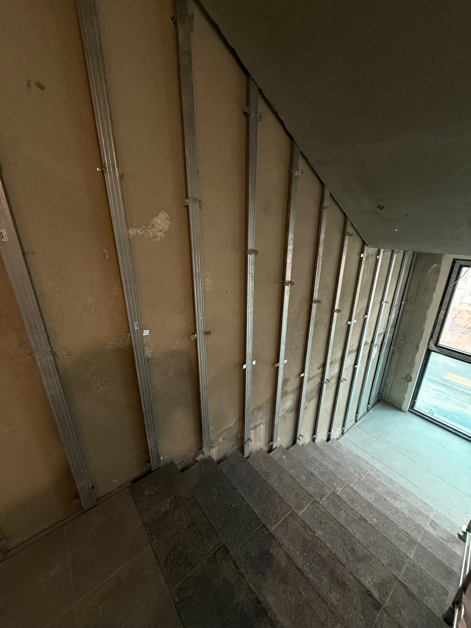 Montaż metalowych profili na ścianie wewnątrz budynku, widoczne schody i okno. Surowy stan wykończenia, przygotowanie do dalszych prac.