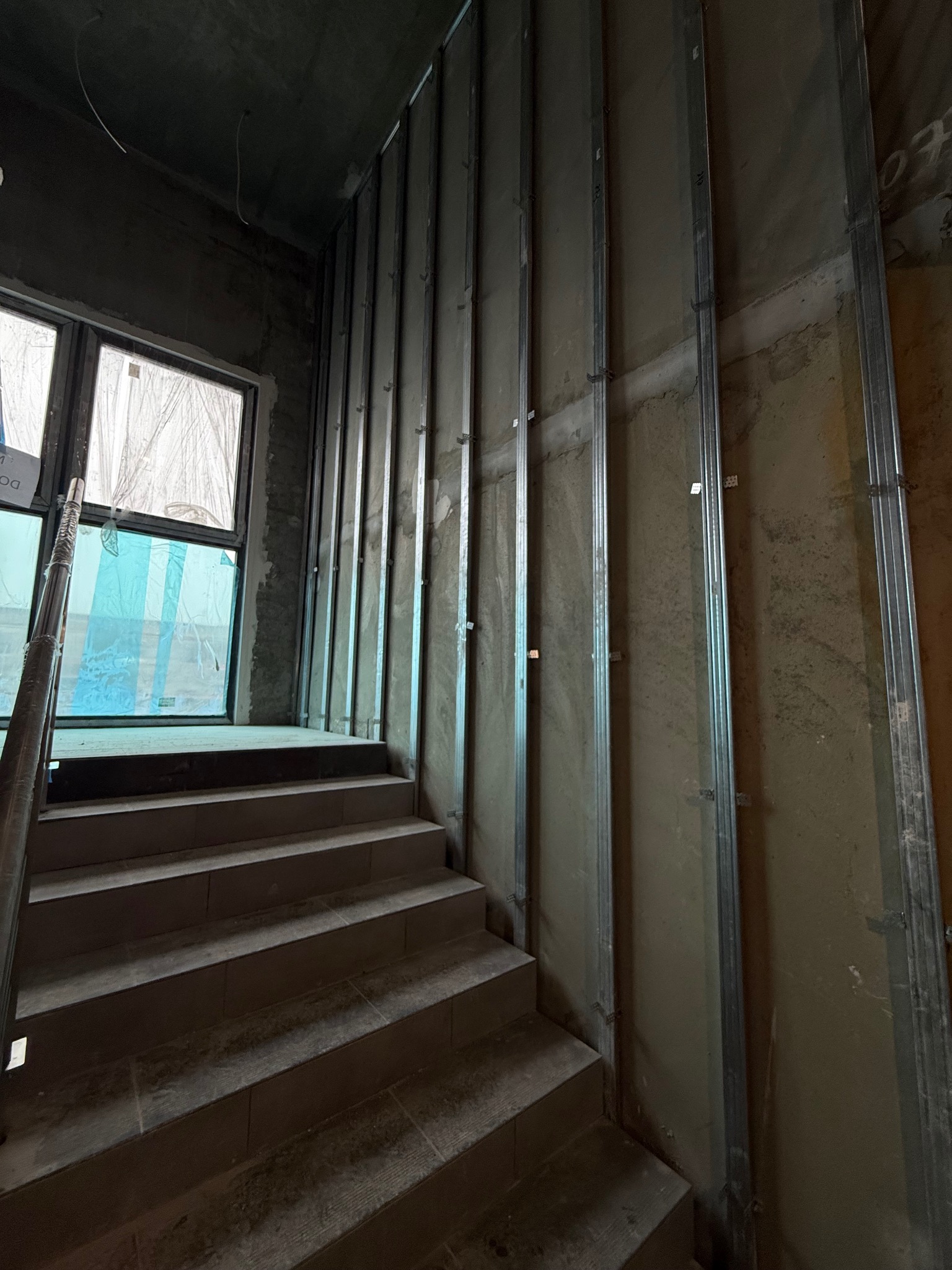 Wnętrze w trakcie wykańczania: schody z płytek prowadzą do okna, obok ściana z metalowymi profilami pionowymi, przygotowana pod zabudowę. Surowy stan, widoczne niedoskonałości.