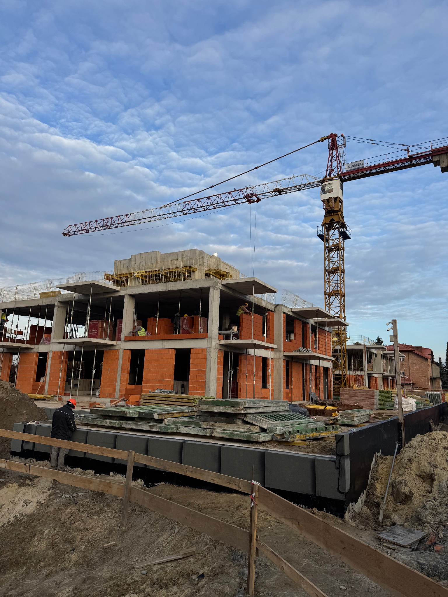 Wznoszenie dwukondygnacyjnego budynku z czerwonej cegły. Widoczny żuraw budowlany, pracownicy w kaskach i zabezpieczone fundamenty. Niebo z chmurami w tle.