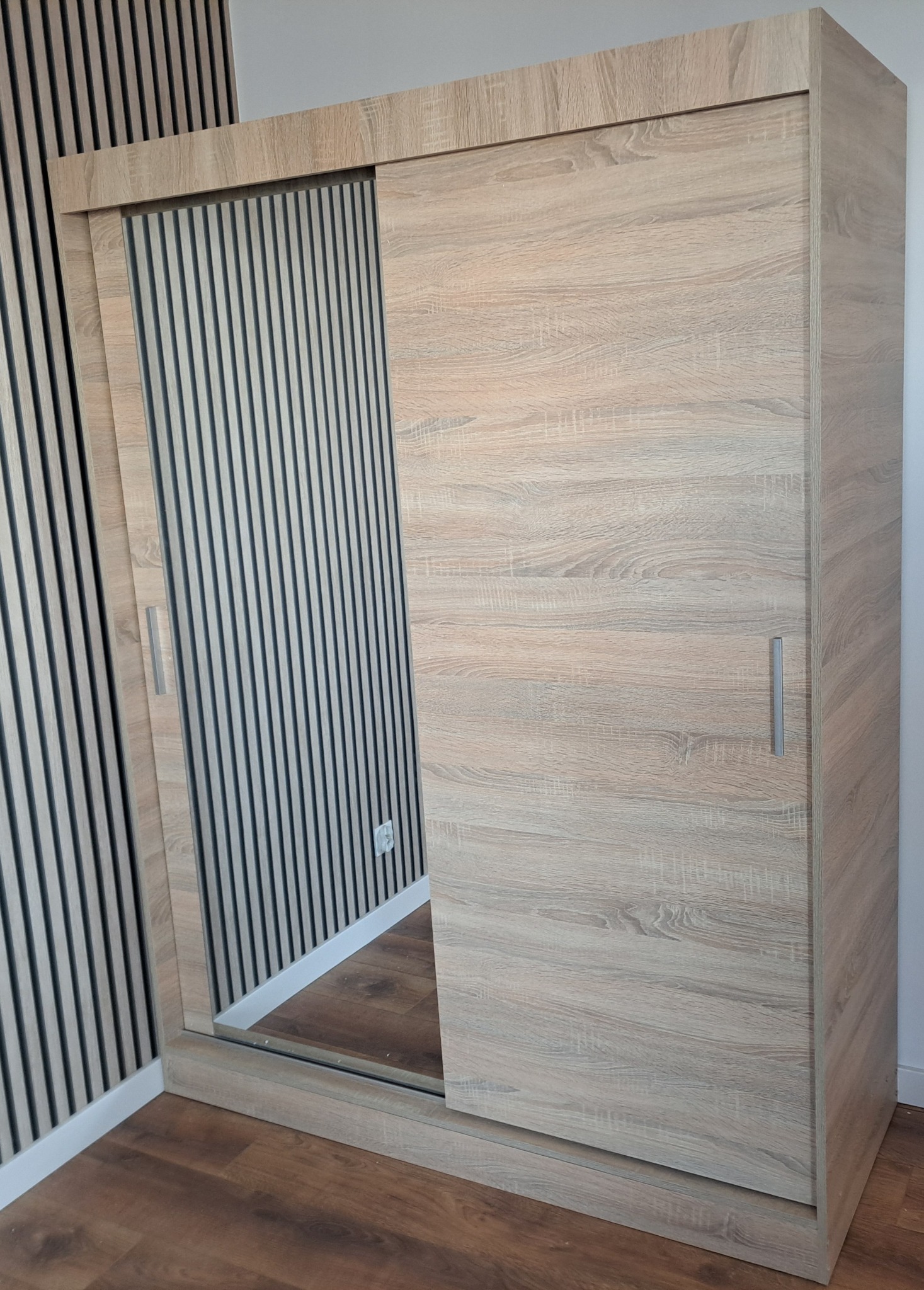 Zamontowana szafa przesuwna z lustrem i wykończeniem w kolorze jasnego drewna, z widoczną ścianą z dekoracyjnymi panelami pionowymi w tle.