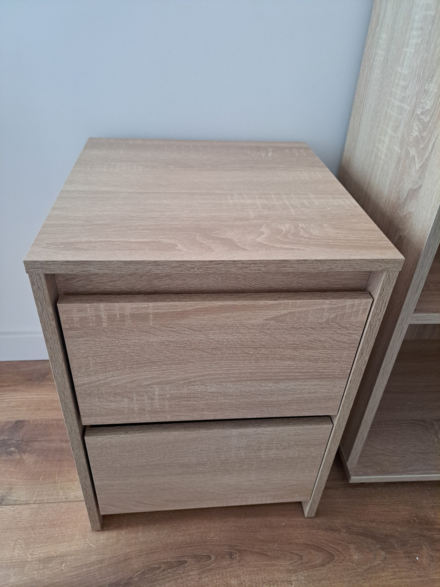 Szafka nocna z dwoma szufladami w odcieniach jasnego drewna, ustawiona na drewnianej podłodze, z fragmentem regału widocznym z boku.