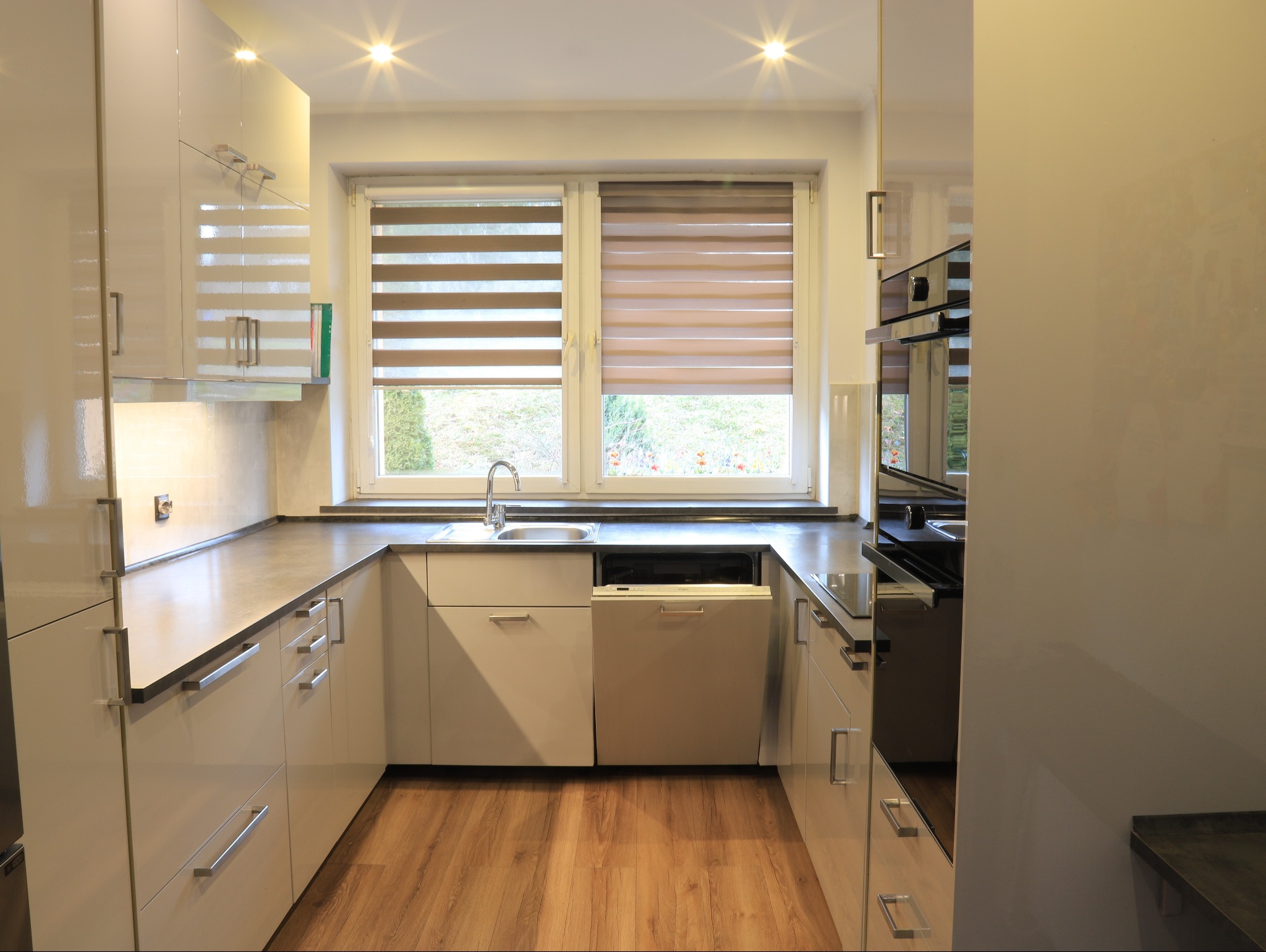 Nowoczesna, jasna kuchnia z białymi, lakierowanymi frontami, szarym blatem i drewnianą podłogą. Widoczny zlew, bateria, okno z roletami dzień-noc i zabudowany piekarnik.