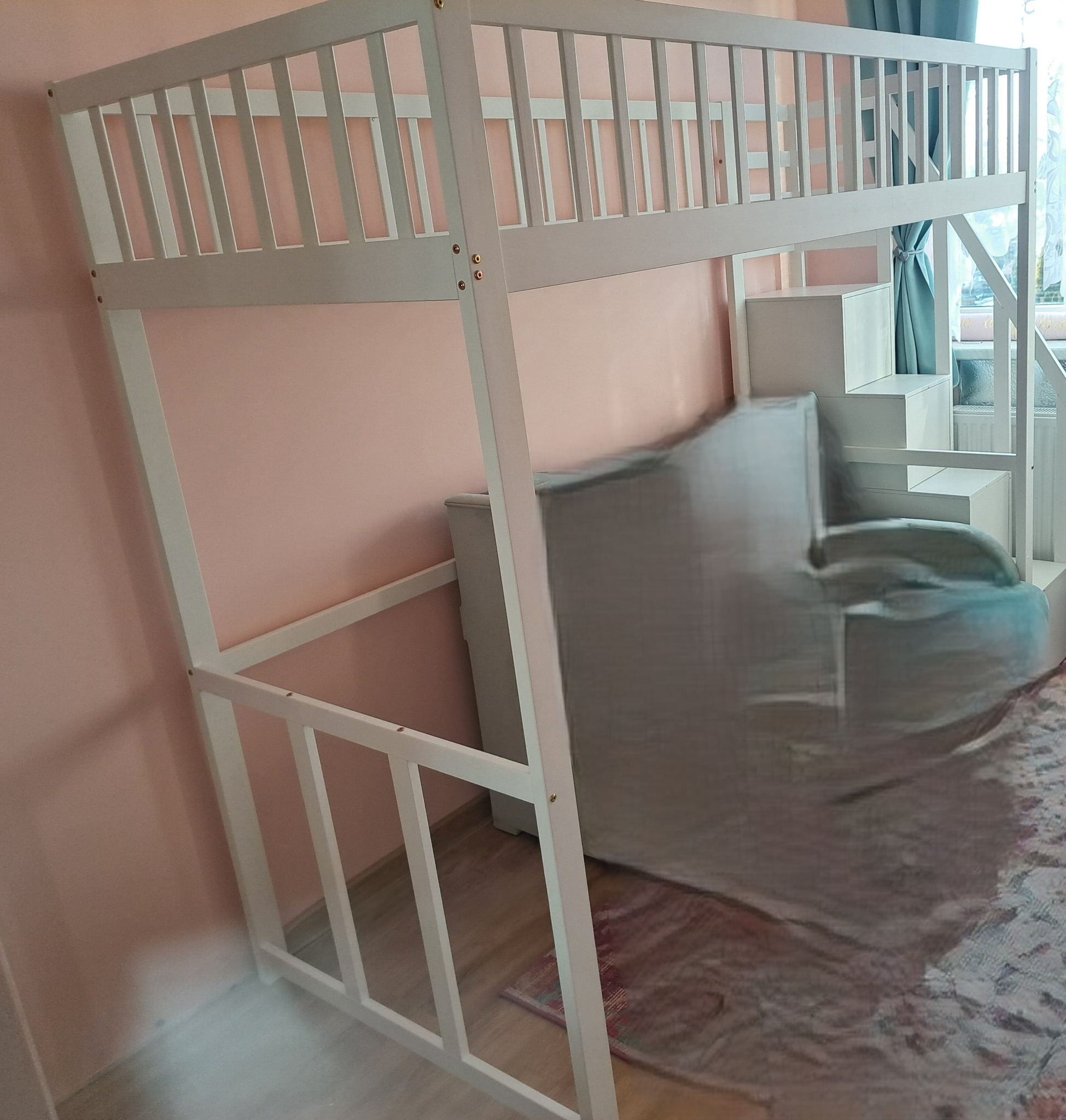 Białe łóżko piętrowe z drabinką schodkową, częściowo zasłonięte przez sofę, na tle różowej ściany i wzorzystego dywanu.