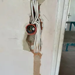 Otwór w ścianie z puszką elektryczną i wystającymi białymi przewodami, widoczne złącza WAGO. Ściana w surowym stanie, obok framuga drzwi. Prace elektryczne w trakcie remontu.