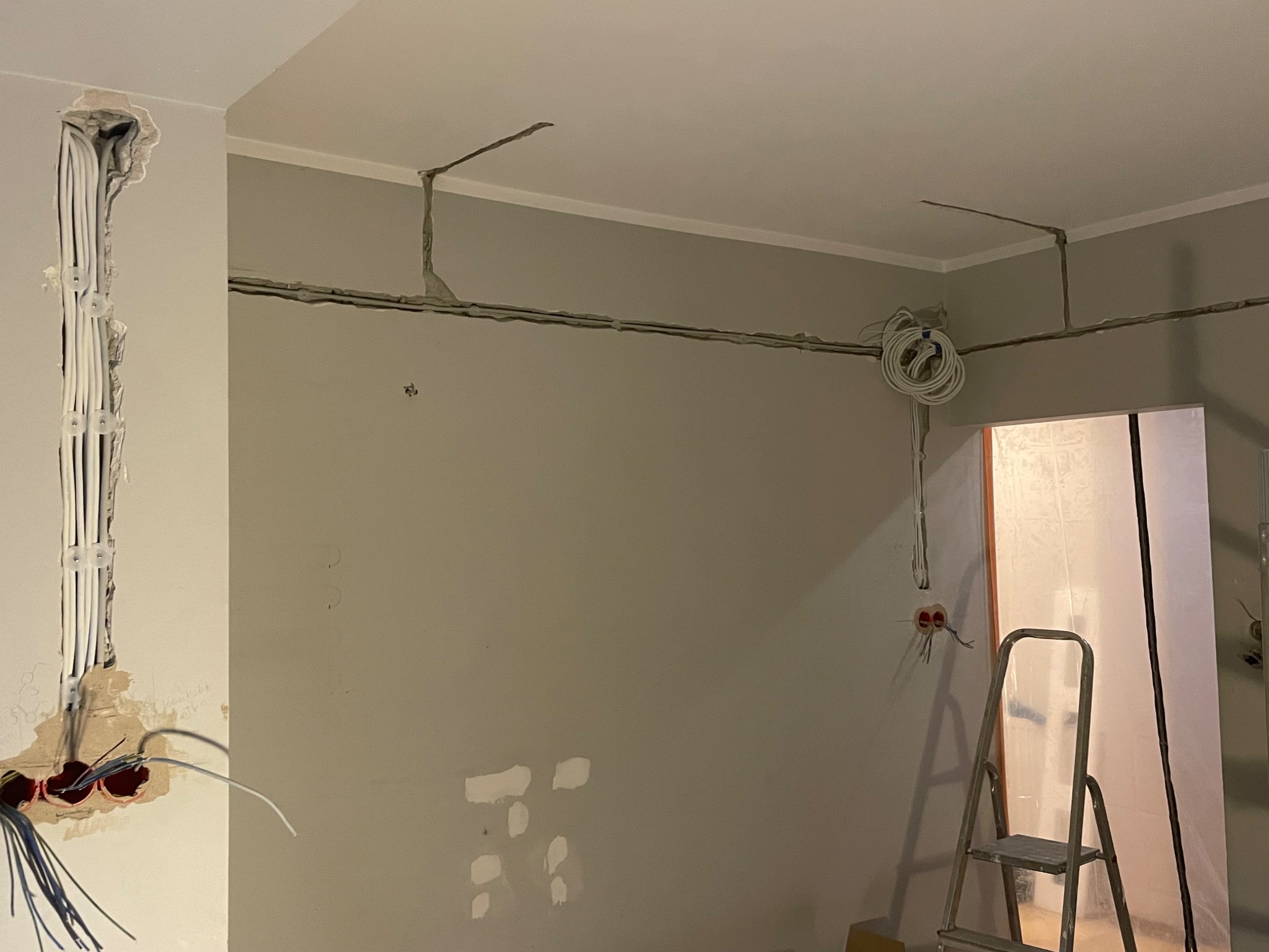 Szare wnętrze w trakcie prac elektrycznych: kable w ścianach i suficie, puszki elektryczne, drabina. Widoczne etapy montażu instalacji.