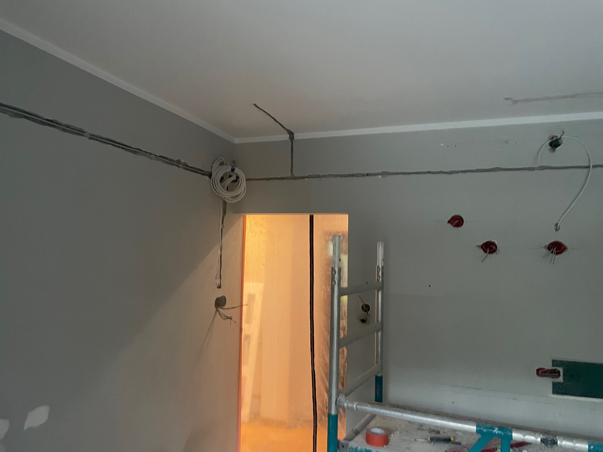 Szare wnętrze w trakcie prac elektrycznych: kable poprowadzone po ścianach, puszki pod gniazdka, drabina malarska. Widok na pomalowane ściany i sufit.