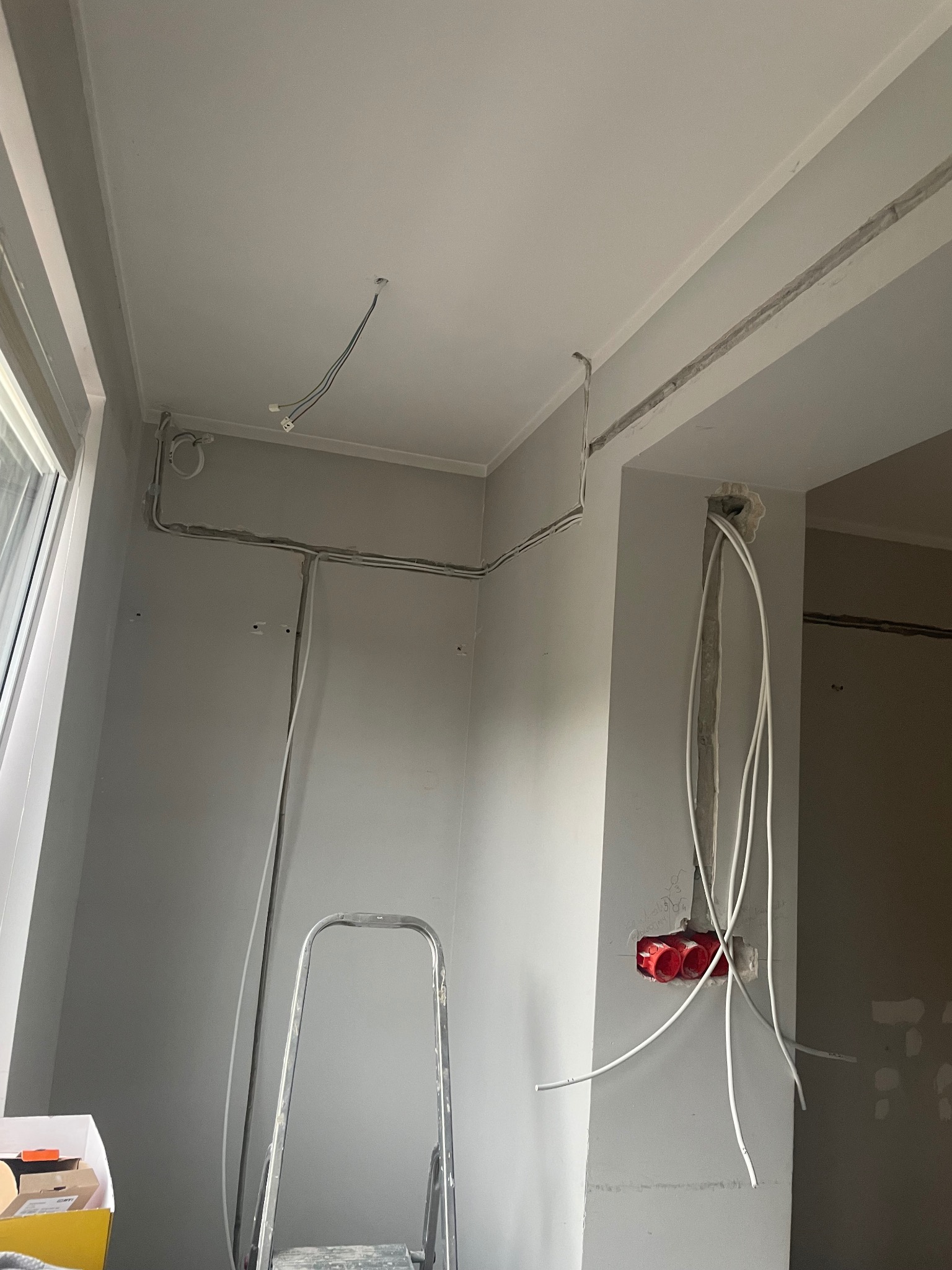 Szare wnętrze w trakcie instalacji elektrycznej: kable prowadzone po ścianach i suficie, czerwone puszki podtynkowe, drabina. Widok na wnętrze z oknem.