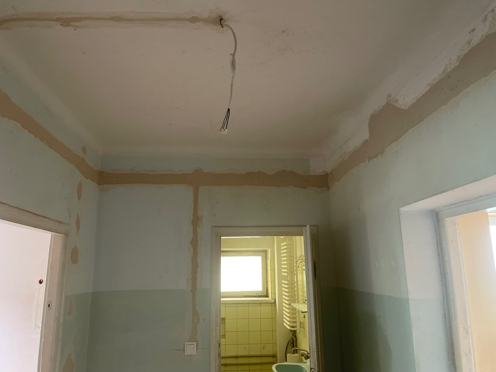 Wnętrze w trakcie remontu: widoczne przygotowane ściany, sufit z wystającym przewodem elektrycznym. Dwa wejścia do innych pomieszczeń. Ujęcie z dołu.
