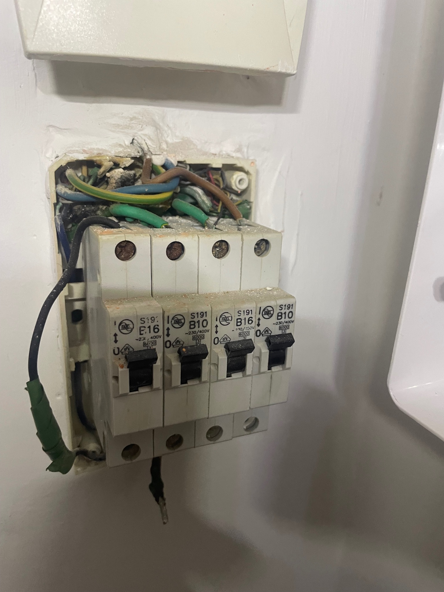 Otwarta skrzynka elektryczna z widocznymi bezpiecznikami i przewodami. Widoczne ślady uszkodzeń i zabrudzeń. Niechlujne wykończenie otworu w ścianie.