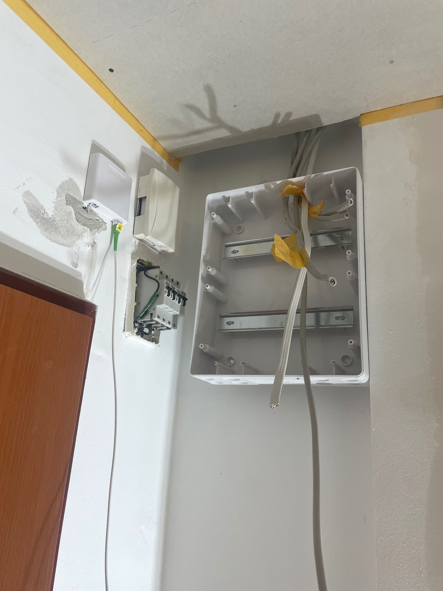 Otwarta skrzynka elektryczna z przewodami wewnątrz, obok gniazdko światłowodowe i puszka z bezpiecznikami. Ściany przygotowane do malowania, zabezpieczone taśmą.