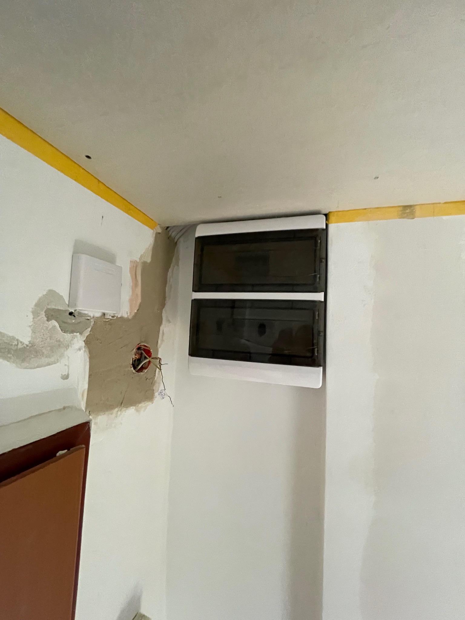 Nowa, dwurzędowa skrzynka bezpiecznikowa zamontowana na ścianie w rogu pomieszczenia, obok puszki elektrycznej z wystającymi przewodami. Ściana przygotowana do malowania.
