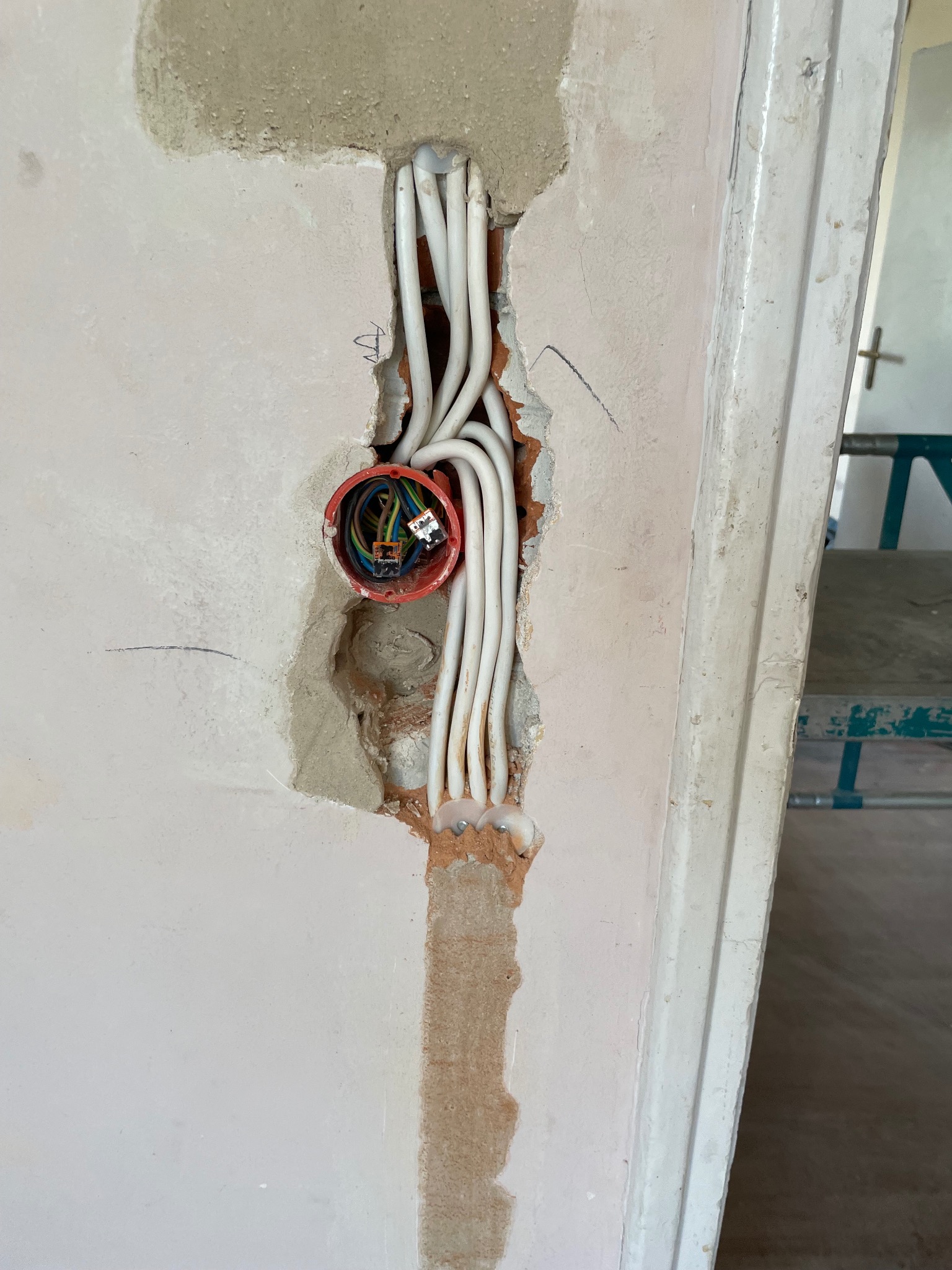 Otwór w ścianie z puszką elektryczną i wystającymi białymi przewodami, widoczne złącza WAGO. Ściana w surowym stanie, obok framuga drzwi. Prace elektryczne w trakcie remontu.