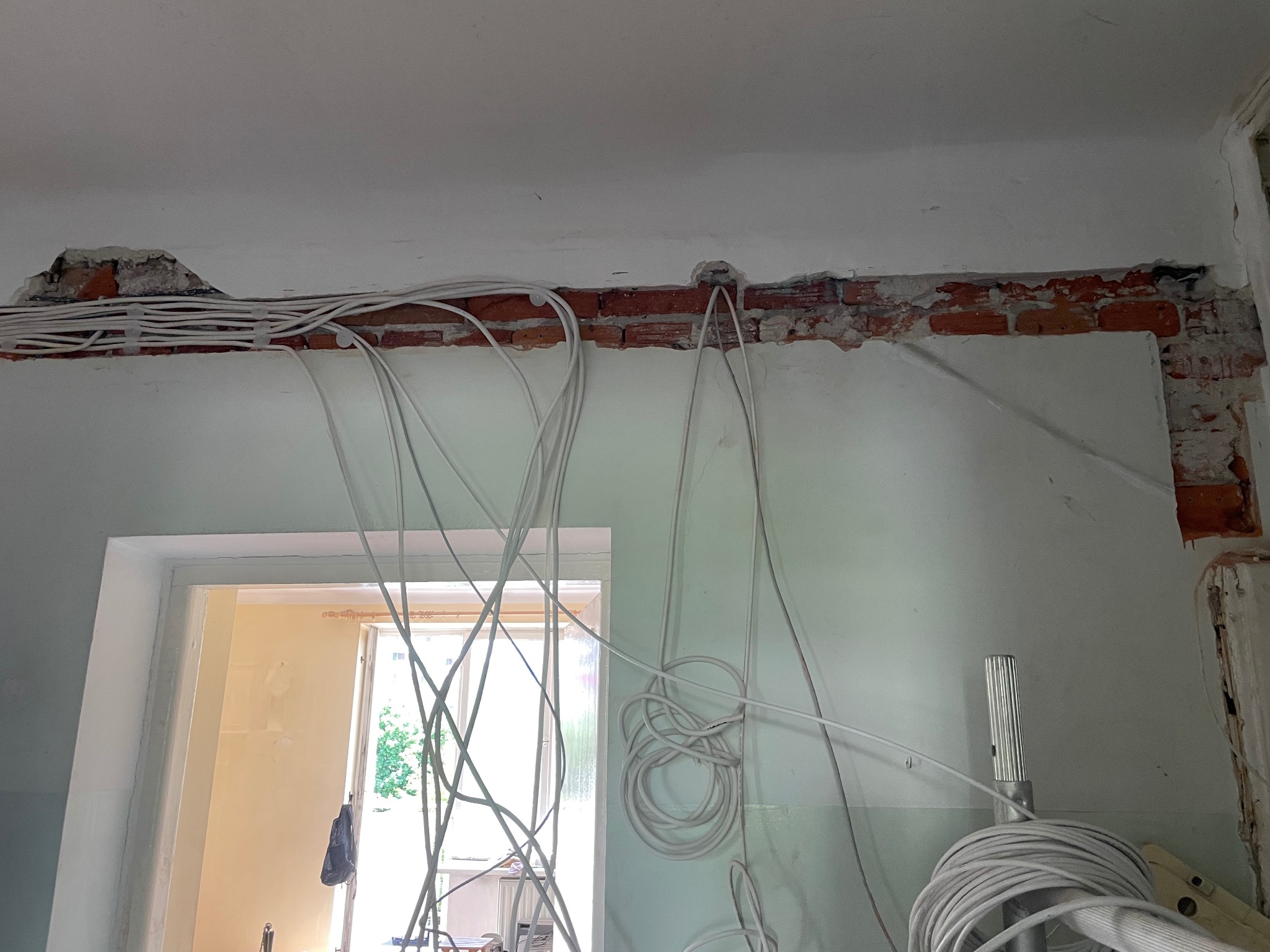Białe kable elektryczne ułożone w bruzdach w ścianie z cegły nad ościeżnicą drzwiową, w trakcie prac instalacyjnych. Widoczne narzędzia.