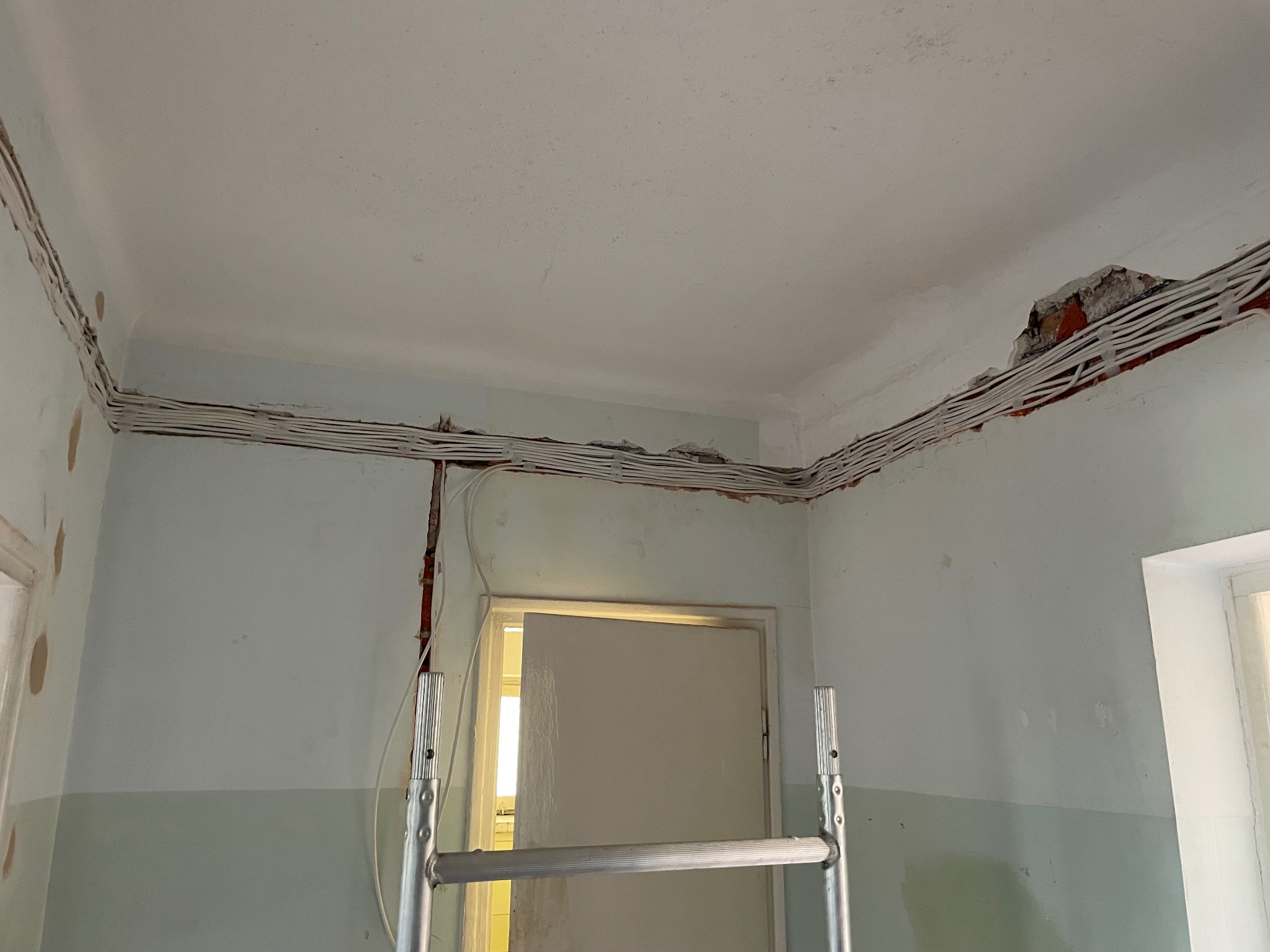 Wnętrze w trakcie remontu z widocznymi kablami elektrycznymi ułożonymi wzdłuż ścian pod sufitem, drabina rozstawiona w pomieszczeniu.