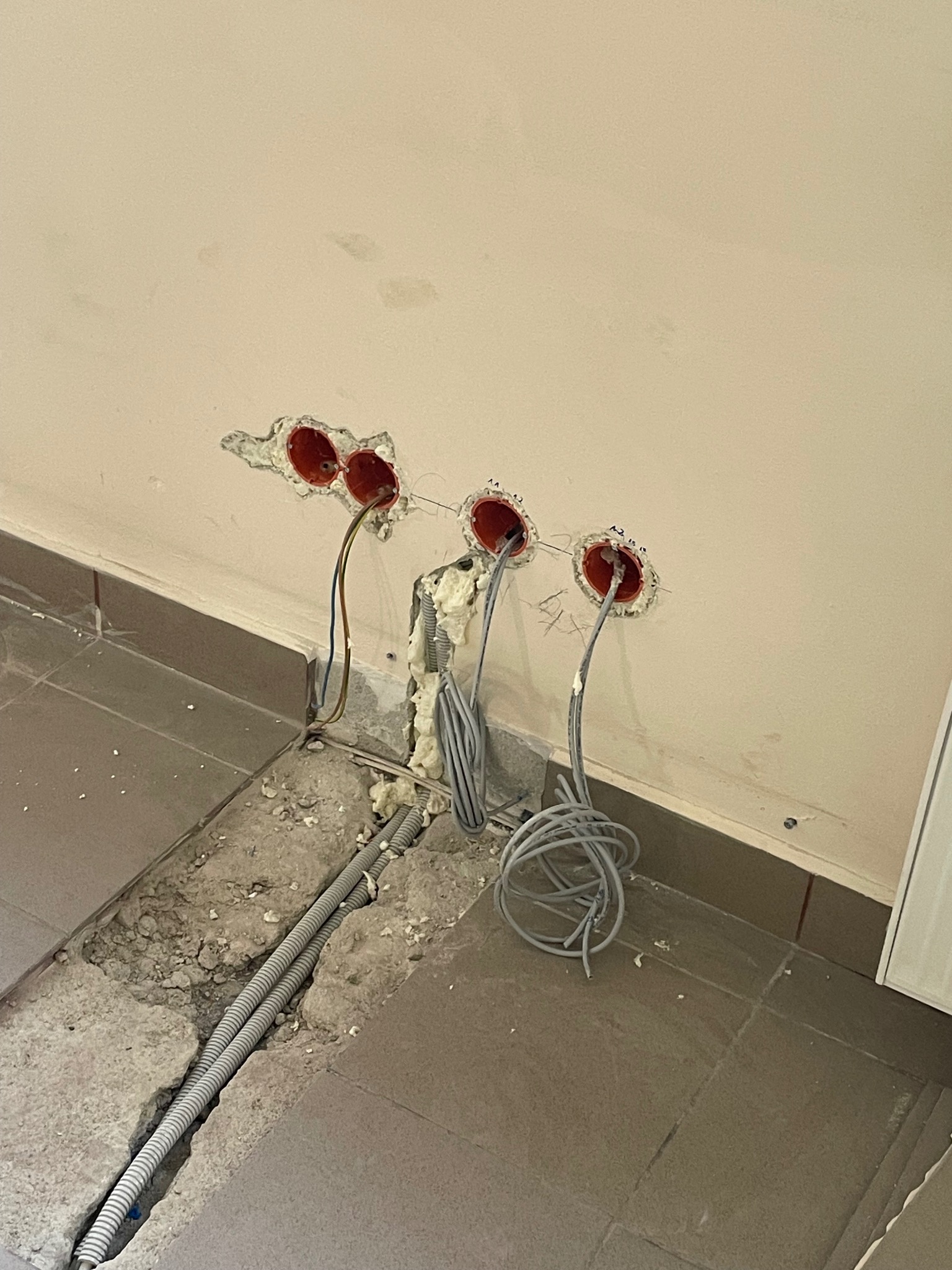 Przygotowane otwory w ścianie z wystającymi przewodami elektrycznymi. Na podłodze kable w peszlach, ślady po kuciu. Widoczne pomarańczowe puszki instalacyjne.