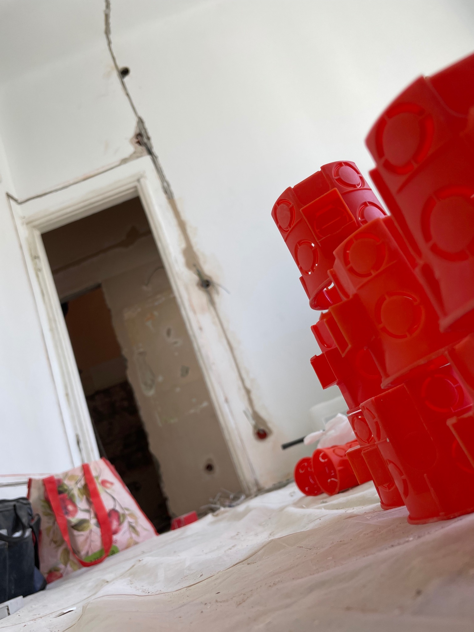 Stos czerwonych puszek elektrycznych na podłodze w trakcie remontu. Widoczne kable w ścianie i torba z motywem owocowym.