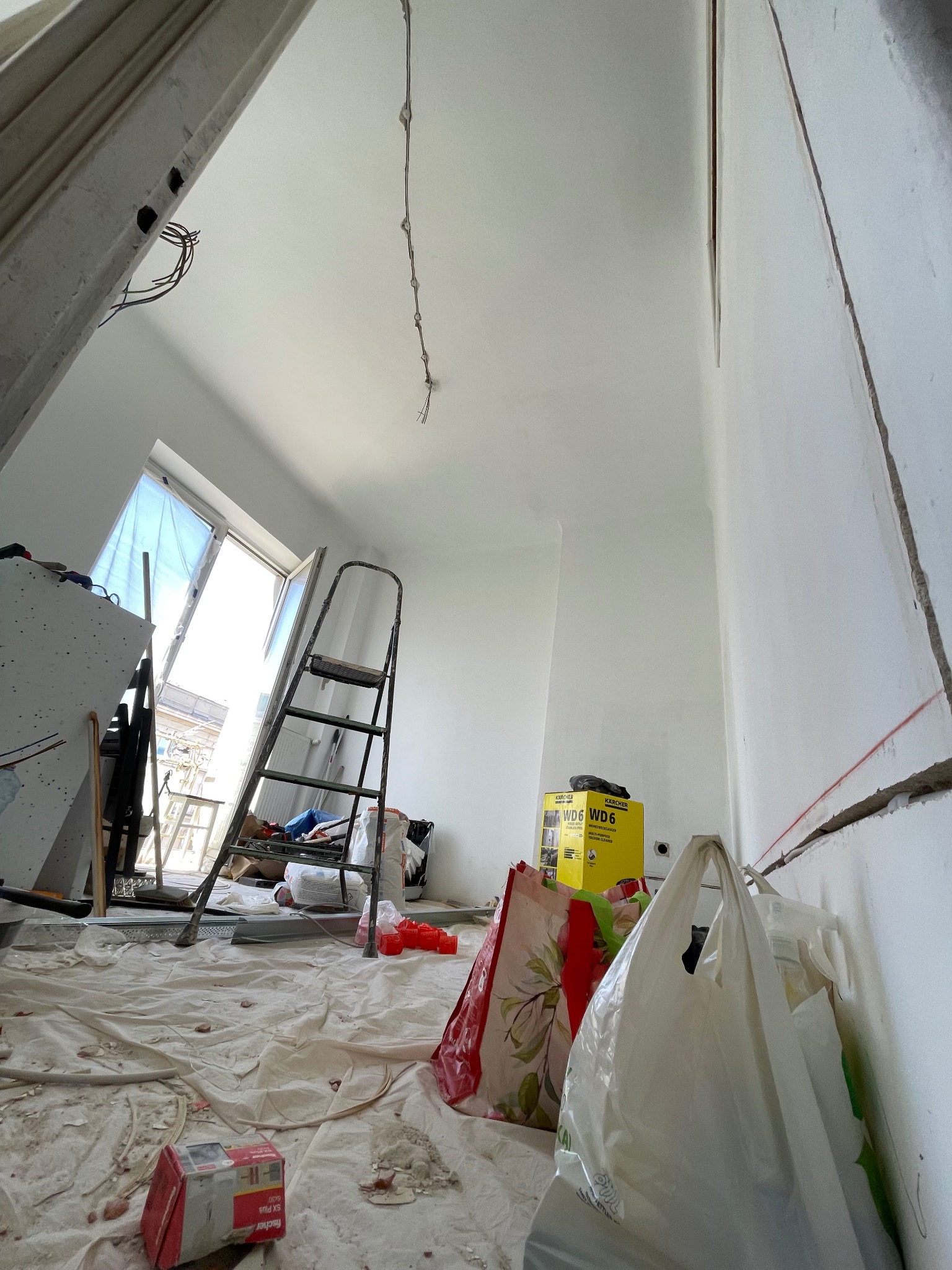 Wnętrze w trakcie prac elektrycznych: wiszące kable, drabina, narzędzia i materiały budowlane. Widoczne bruzdy w ścianie pod instalację elektryczną.
