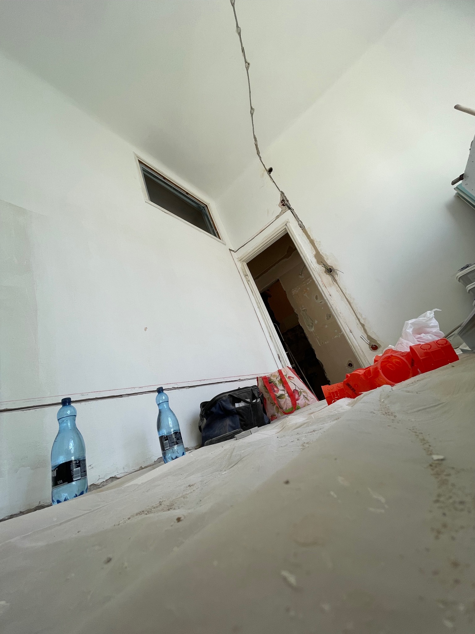 Remontowany pokój z widocznymi przewodami elektrycznymi, zabezpieczoną podłogą, butelkami wody i pomarańczowymi elementami instalacji elektrycznej.