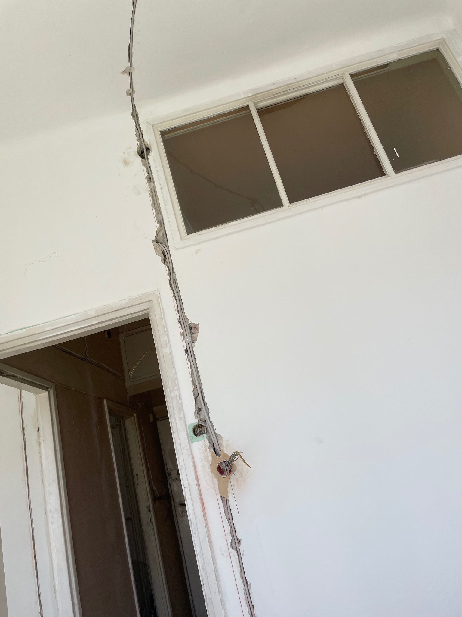 Biała ściana z oknem i wystającymi przewodami elektrycznymi w korycie. Widoczne gniazdko elektryczne i fragmenty instalacji w ścianie. Remont wnętrza.