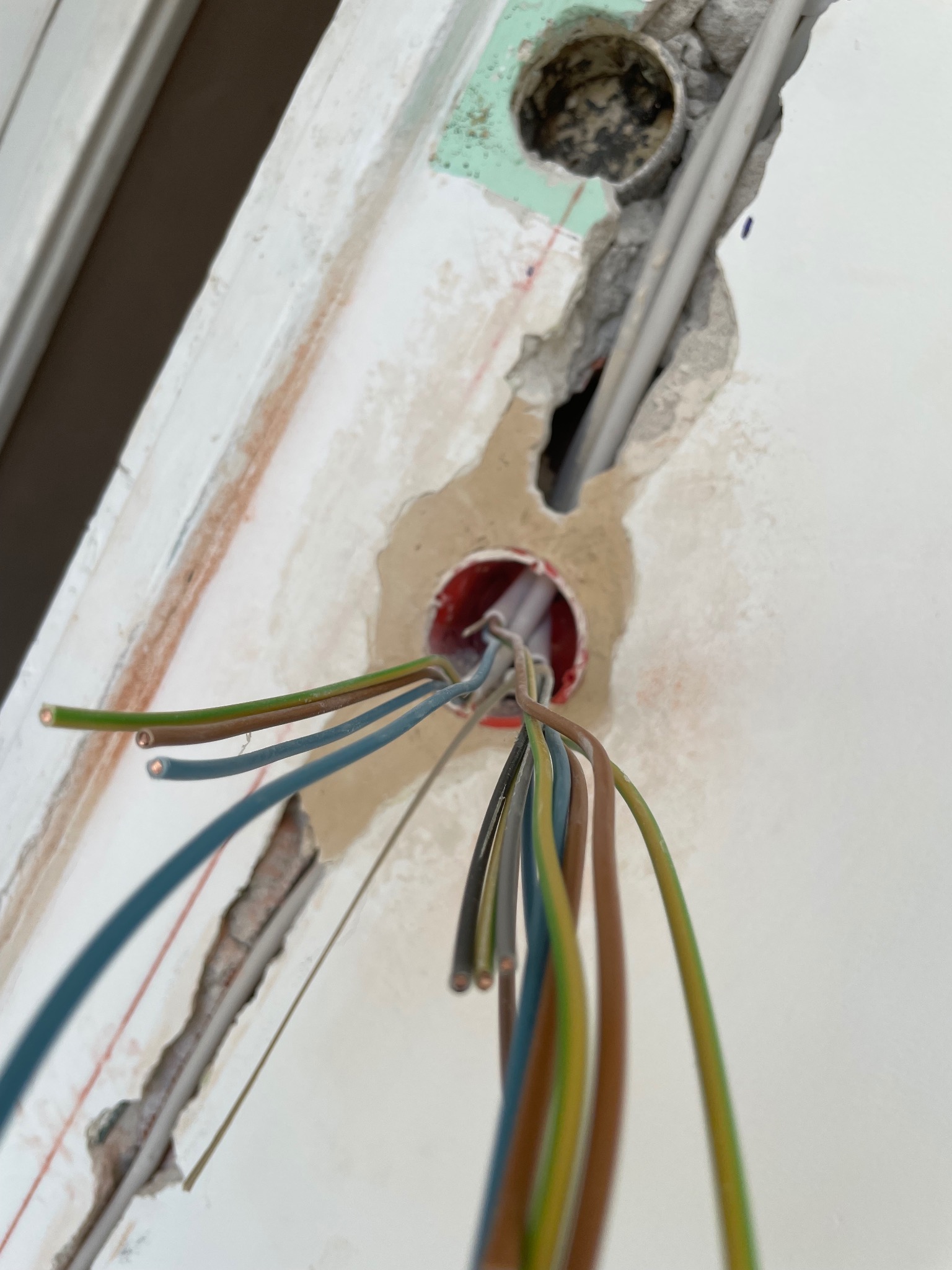 Wystające przewody elektryczne z puszki w ścianie, widoczne bruzdy z kablami w tynku, surowy stan instalacji elektrycznej w trakcie prac wykończeniowych.