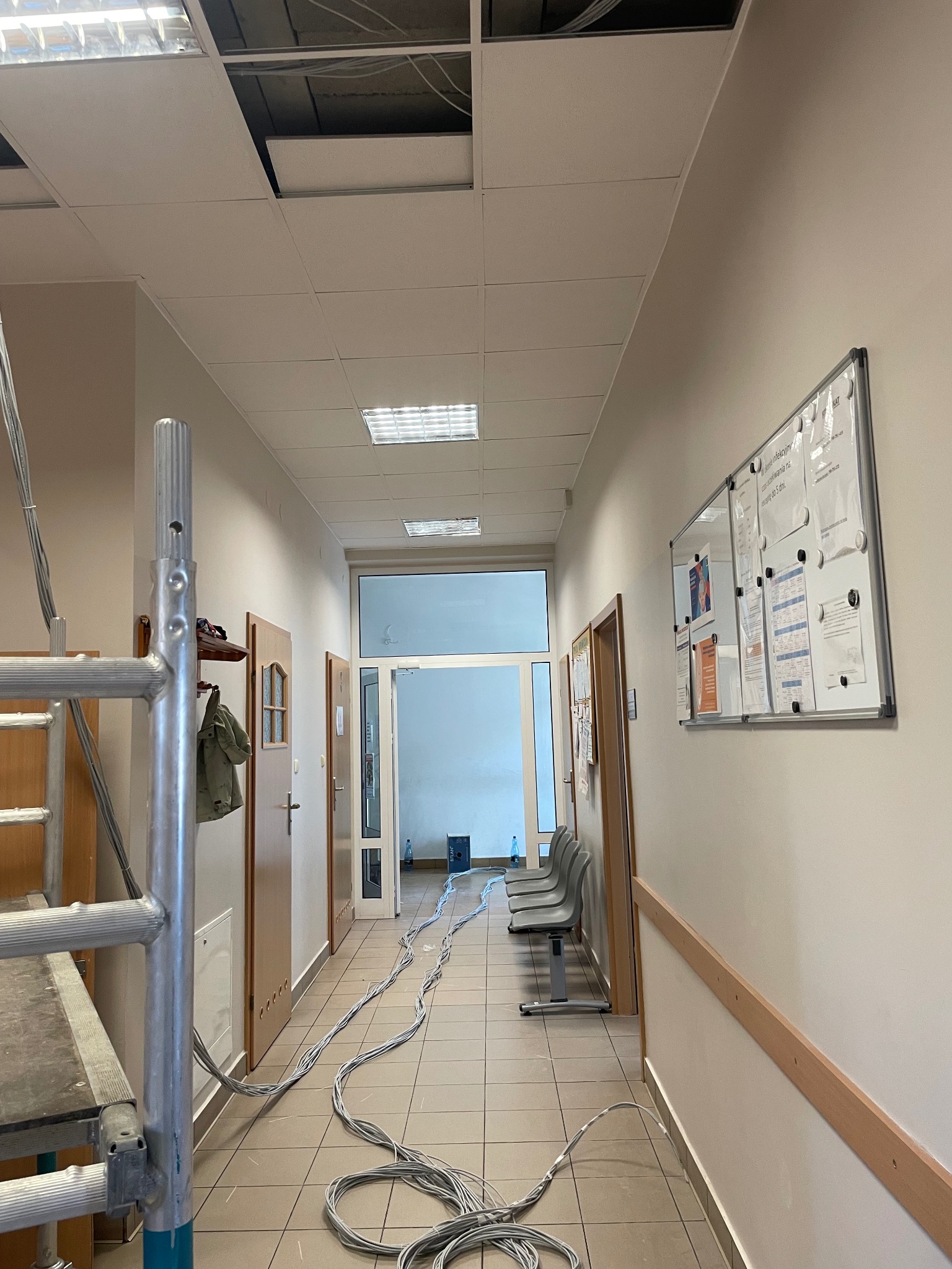 Kable elektryczne rozciągnięte wzdłuż korytarza z częściowo zdjętym sufitem podwieszanym, widoczne rusztowanie i tablica informacyjna na ścianie.
