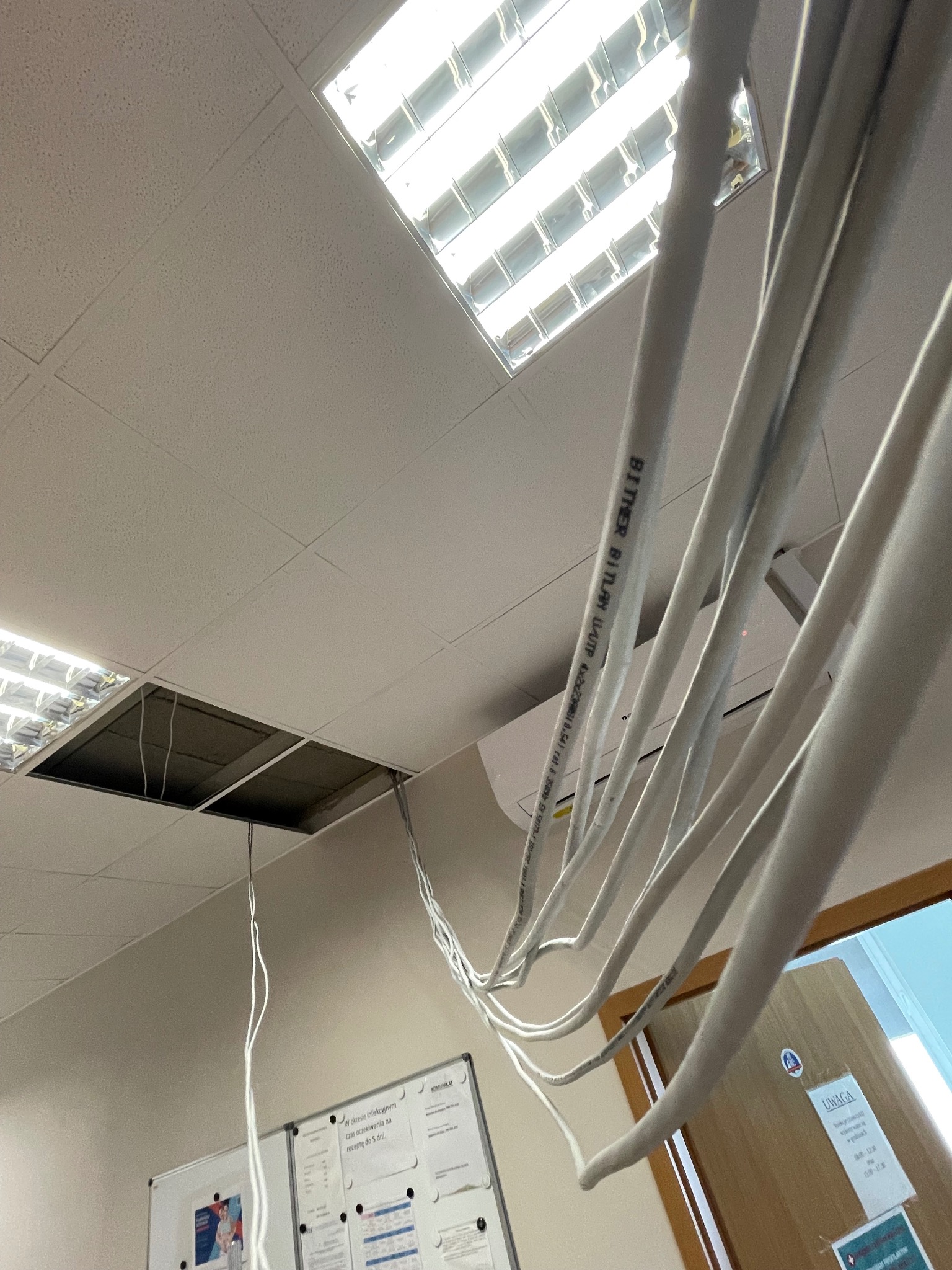 Przewody elektryczne zwisające z sufitu podwieszanego, widoczna oprawa oświetleniowa i klimatyzator. Widok z dołu na instalację w trakcie prac.
