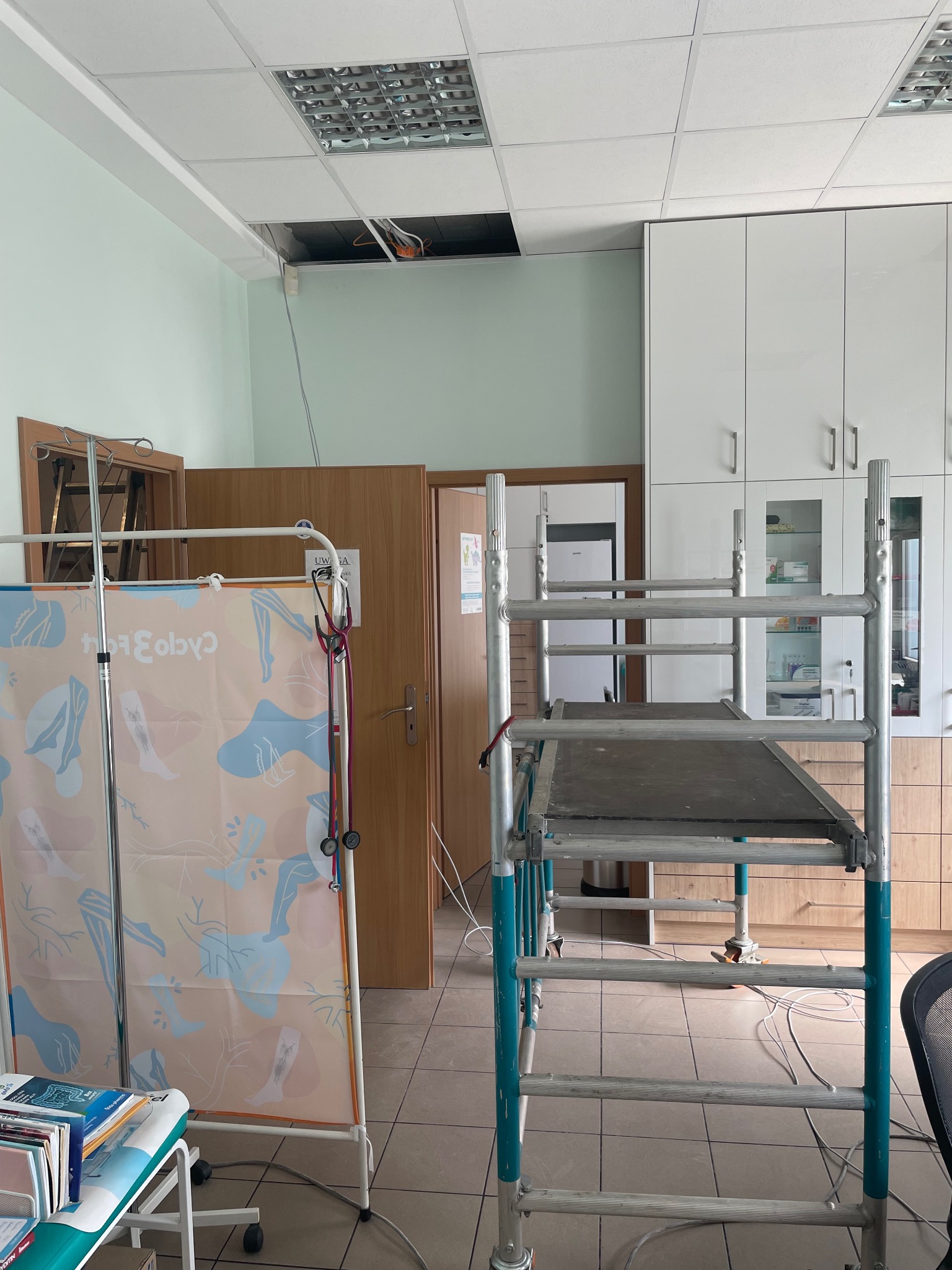 Prace elektryczne w pomieszczeniu: rusztowanie przy dziurze w suficie, widoczne kable. Białe szafki w tle. Instalacja elektryczna w trakcie naprawy lub montażu.