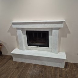 Mobilne Usługi Kamieniarskie JFM- Stone Łukasz Wierzbicki - Biały, marmurowy kominek z czarnym wkładem, na tle beżowej ściany i brązowej podłogi imitującej drewno. Widok z przodu, centralne ujęcie.