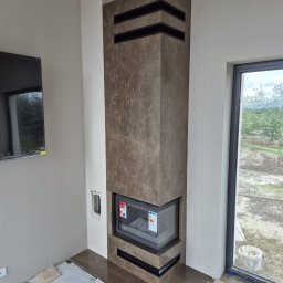 Mobilne Usługi Kamieniarskie JFM- Stone Łukasz Wierzbicki - Nowoczesny kominek obłożony brązowym kamieniem z czarnymi akcentami, w trakcie montażu. Widoczne zabezpieczenia podłogi i fragment okna z widokiem na zewnątrz.