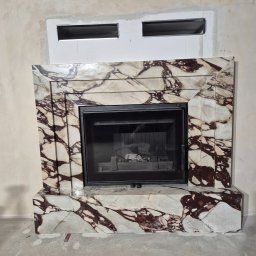 Mobilne Usługi Kamieniarskie JFM- Stone Łukasz Wierzbicki - Marmurowa obudowa kominka z czarnym wkładem, widoczna na tle surowej ściany z przygotowanym otworem wentylacyjnym. Wnętrze kominka z resztkami popiołu.