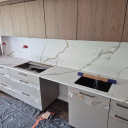 Mobilne Usługi Kamieniarskie JFM- Stone Łukasz Wierzbicki - Jasny blat kuchenny z marmurowym wzorem, zamontowany w nowoczesnej kuchni z szarymi szafkami i drewnianymi górnymi szafkami. Widoczne przygotowania do montażu.