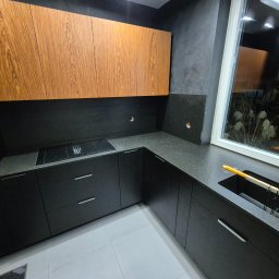 JFM Stone - Czarna kuchnia z drewnianymi szafkami wiszącymi i blatem z ciemnego kamienia w Puszczy Mariańskiej. Nowoczesny design, minimalistyczne uchwyty, okno z widokiem na zieleń.
