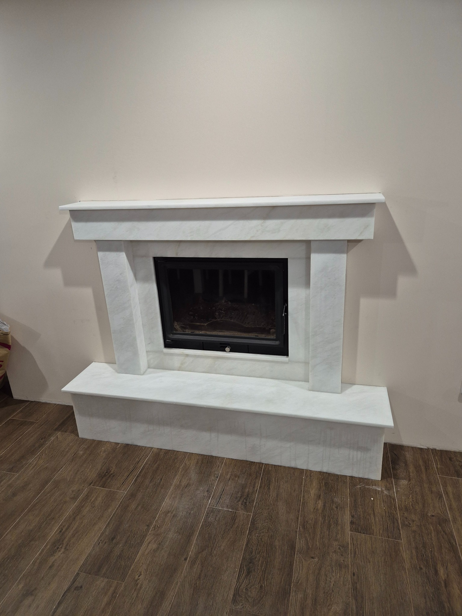 Biały, marmurowy kominek z czarnym wkładem, na tle beżowej ściany i brązowej podłogi imitującej drewno. Widok z przodu, centralne ujęcie.