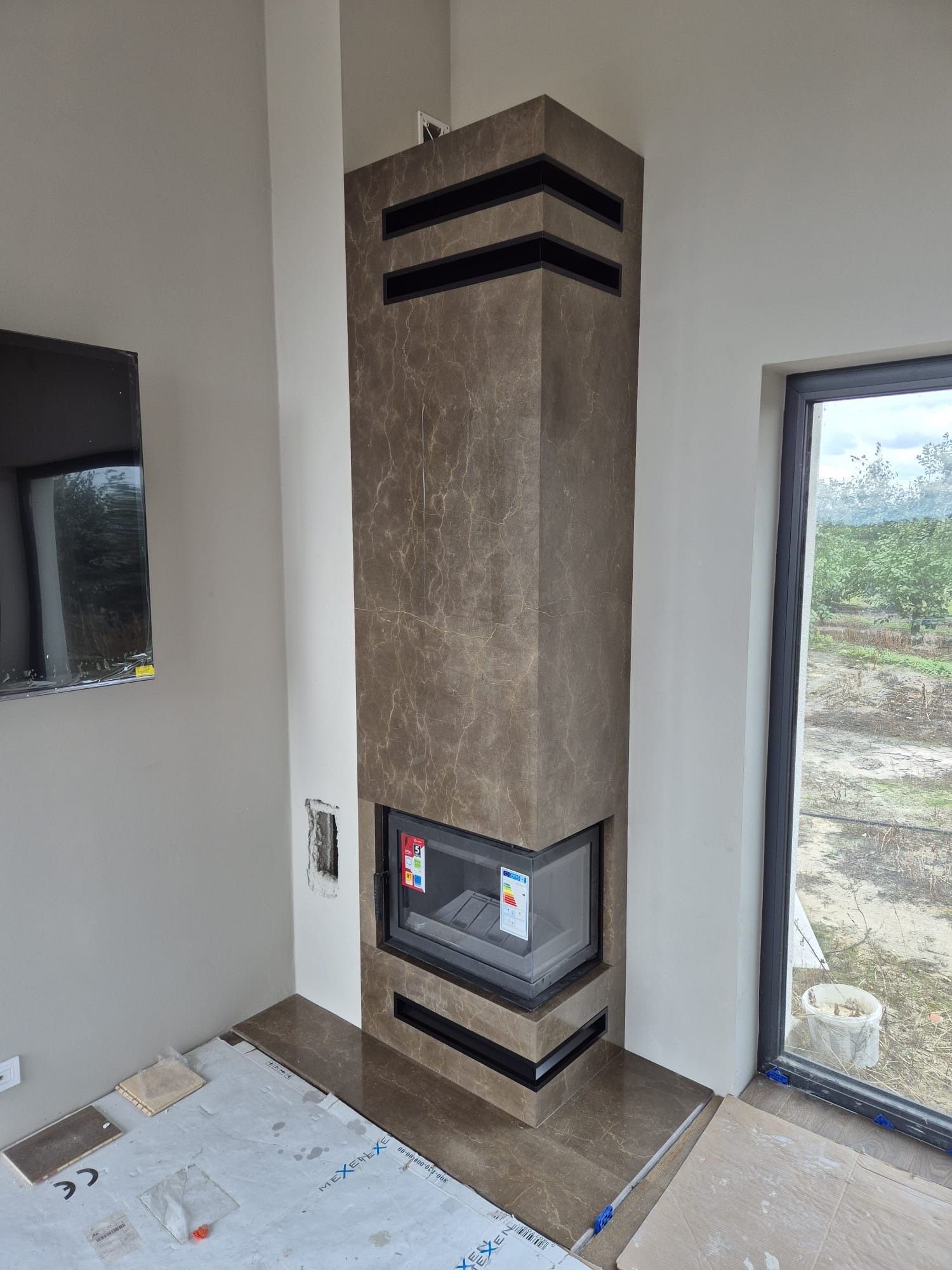 Nowoczesny kominek obłożony brązowym kamieniem z czarnymi akcentami, w trakcie montażu. Widoczne zabezpieczenia podłogi i fragment okna z widokiem na zewnątrz.