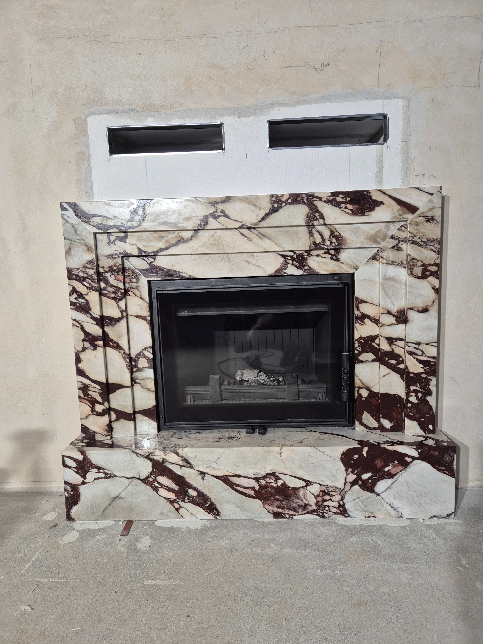 Marmurowa obudowa kominka z czarnym wkładem, widoczna na tle surowej ściany z przygotowanym otworem wentylacyjnym. Wnętrze kominka z resztkami popiołu.