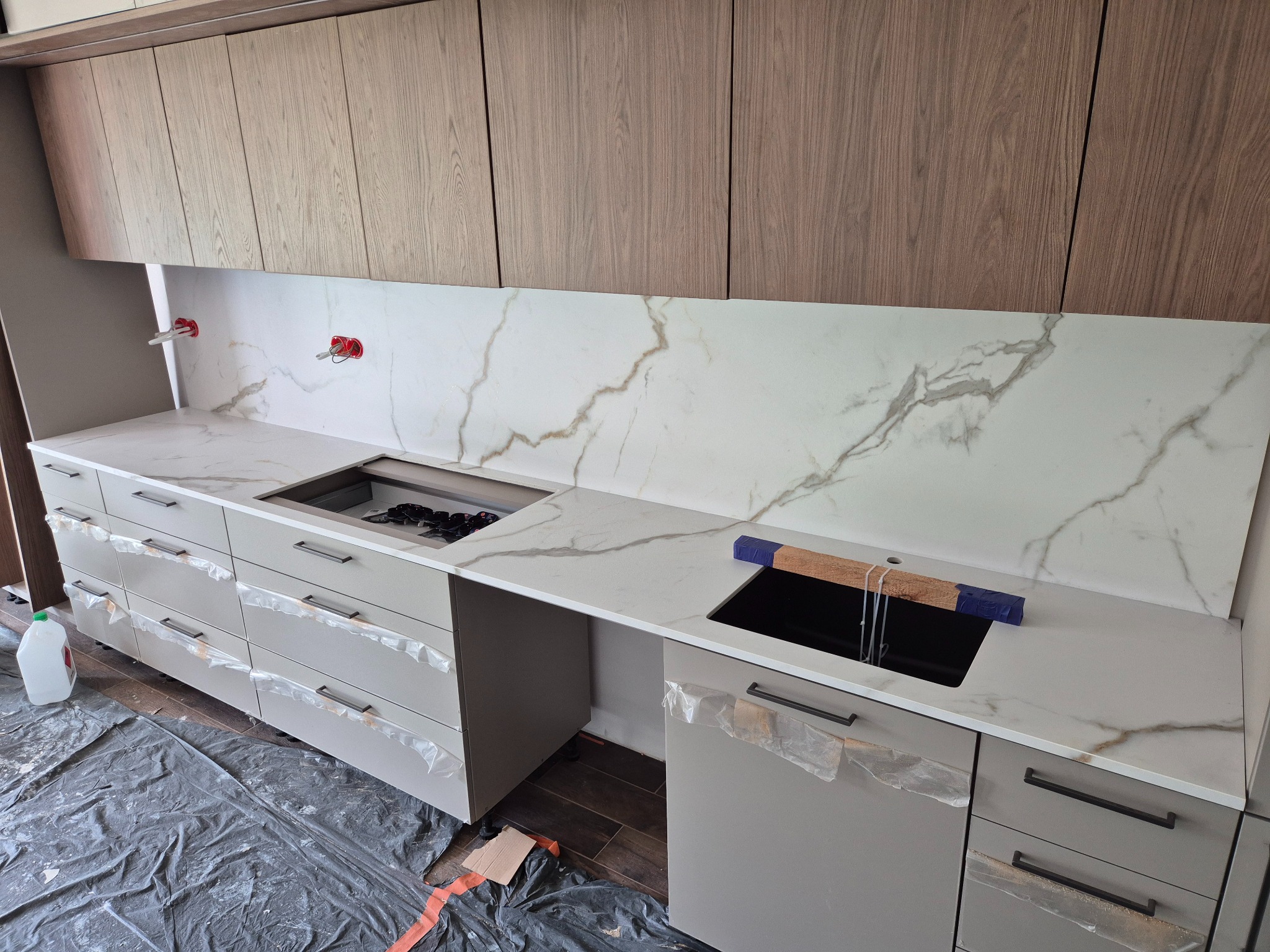 Jasny blat kuchenny z marmurowym wzorem, zamontowany w nowoczesnej kuchni z szarymi szafkami i drewnianymi górnymi szafkami. Widoczne przygotowania do montażu.