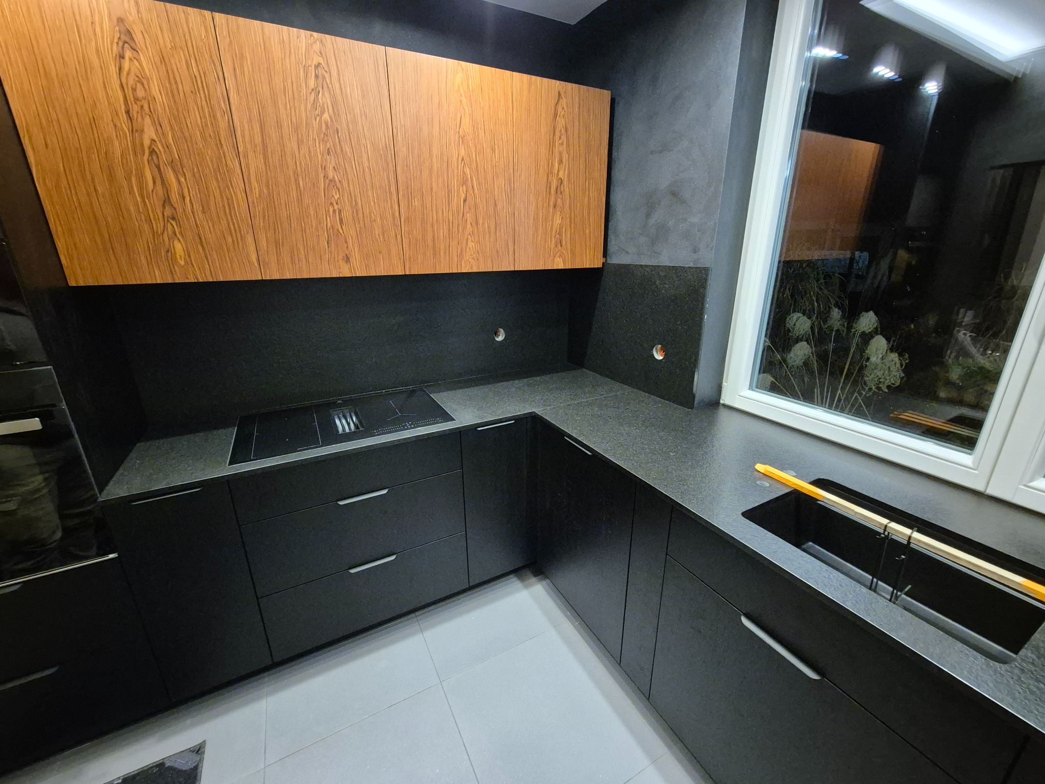 Czarna kuchnia z drewnianymi szafkami wiszącymi i blatem z ciemnego kamienia w Puszczy Mariańskiej. Nowoczesny design, minimalistyczne uchwyty, okno z widokiem na zieleń.
