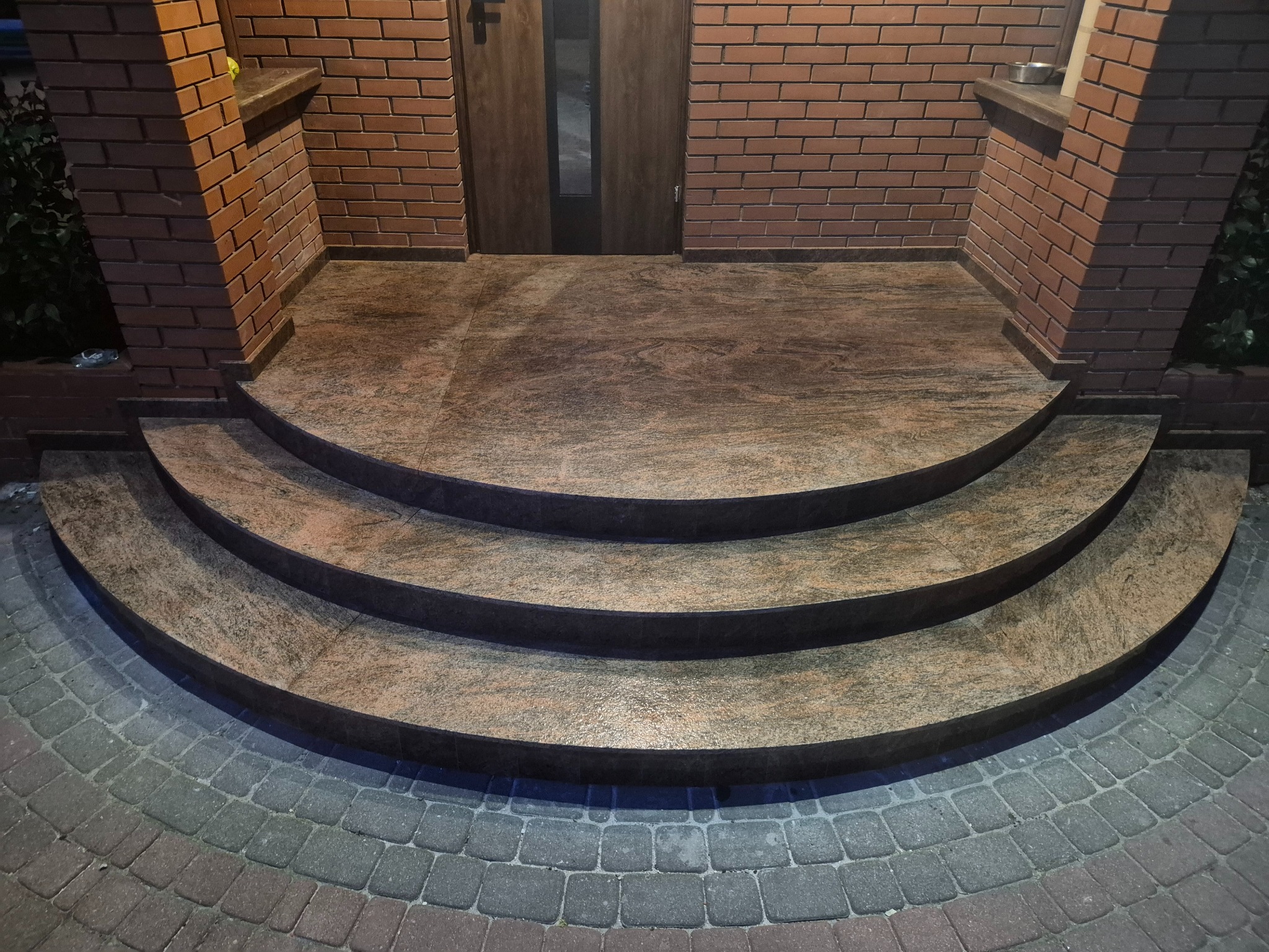 Granitowe schody wejściowe z trzema stopniami, prowadzące do drzwi wejściowych. Czerwona cegła na ścianach i kolumnach. Brukowana ścieżka z kostki.